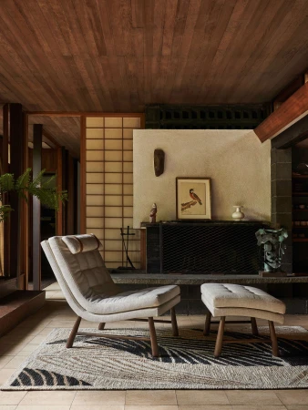 Interior view of a neutral-toned modern living room with a lounge chair, footstool, graphic rug, and decorative fireplace.