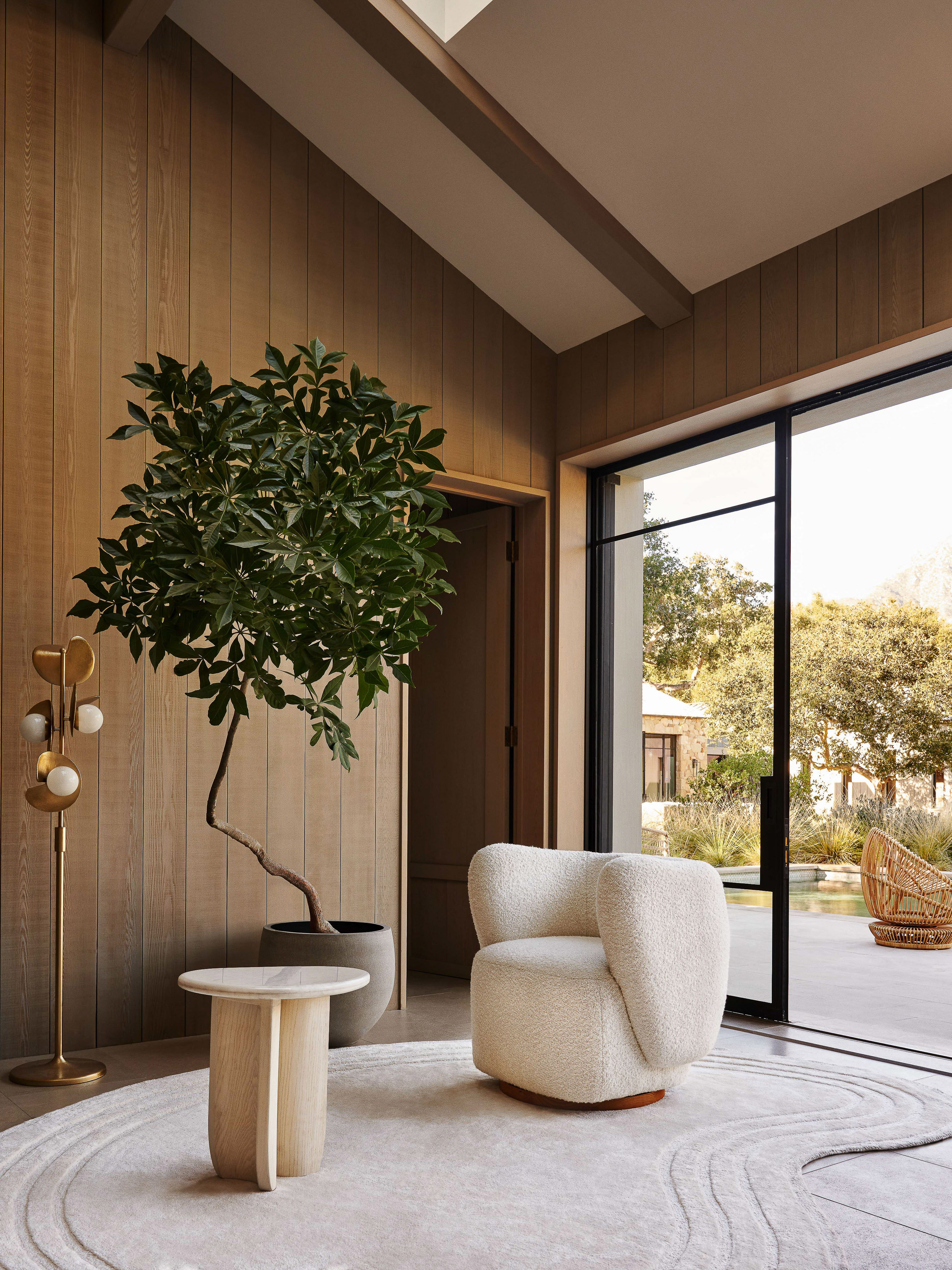 Interior view of a living room with a cream armchair, round coffee table, indoor tree, floor lamp, and neutral patterned rug.