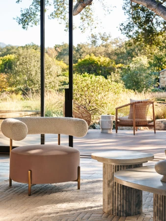 Indoor living room with modern furniture including a velvet-upholstered accent chair, stone coffee table, and a view to outdoor greenery.