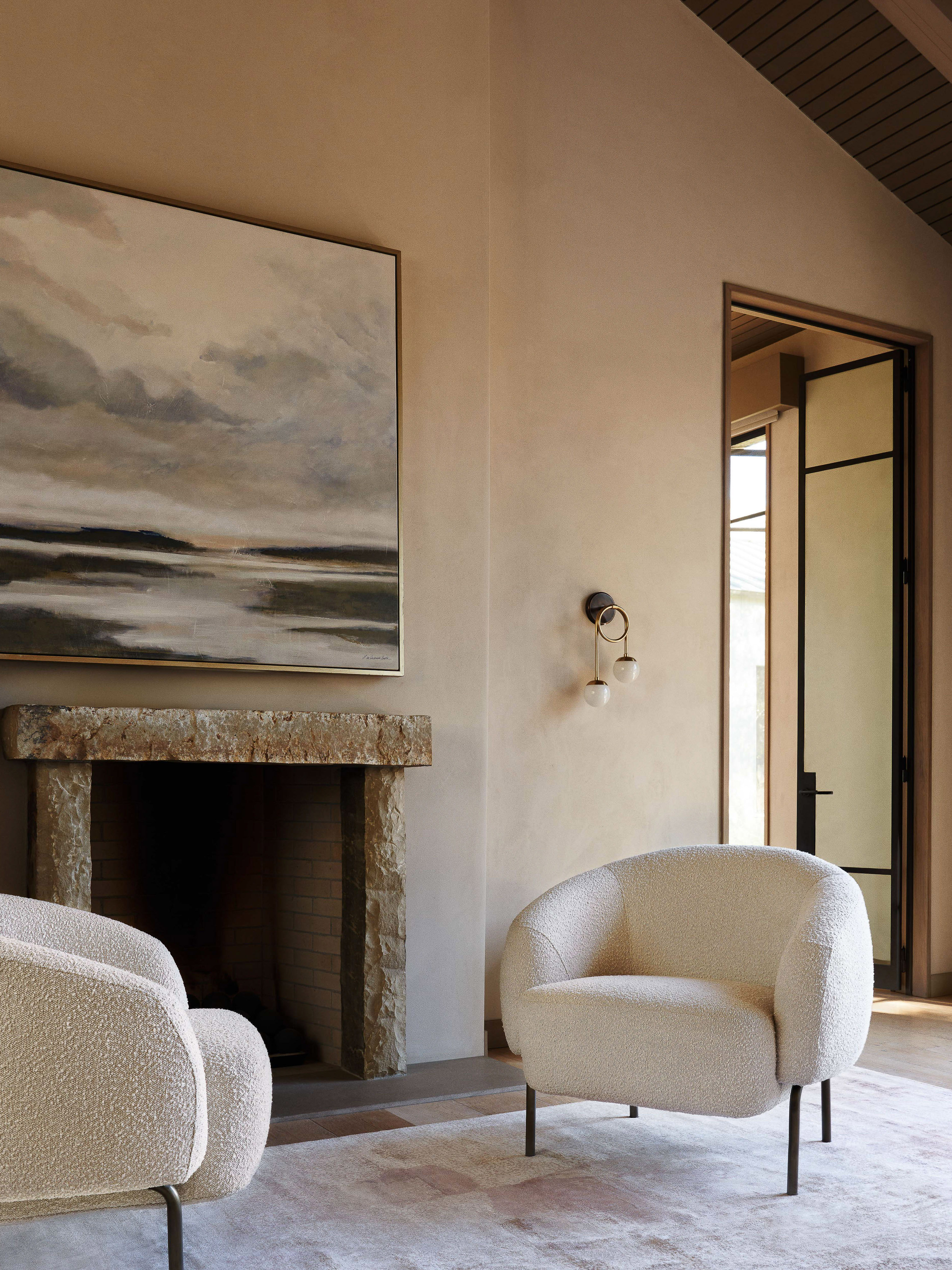 Living room interior with two boucle armchairs, stone fireplace, and abstract painting on a beige wall, creating a cozy atmosphere.