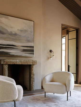 Living room interior with two boucle armchairs, stone fireplace, and abstract painting on a beige wall, creating a cozy atmosphere.