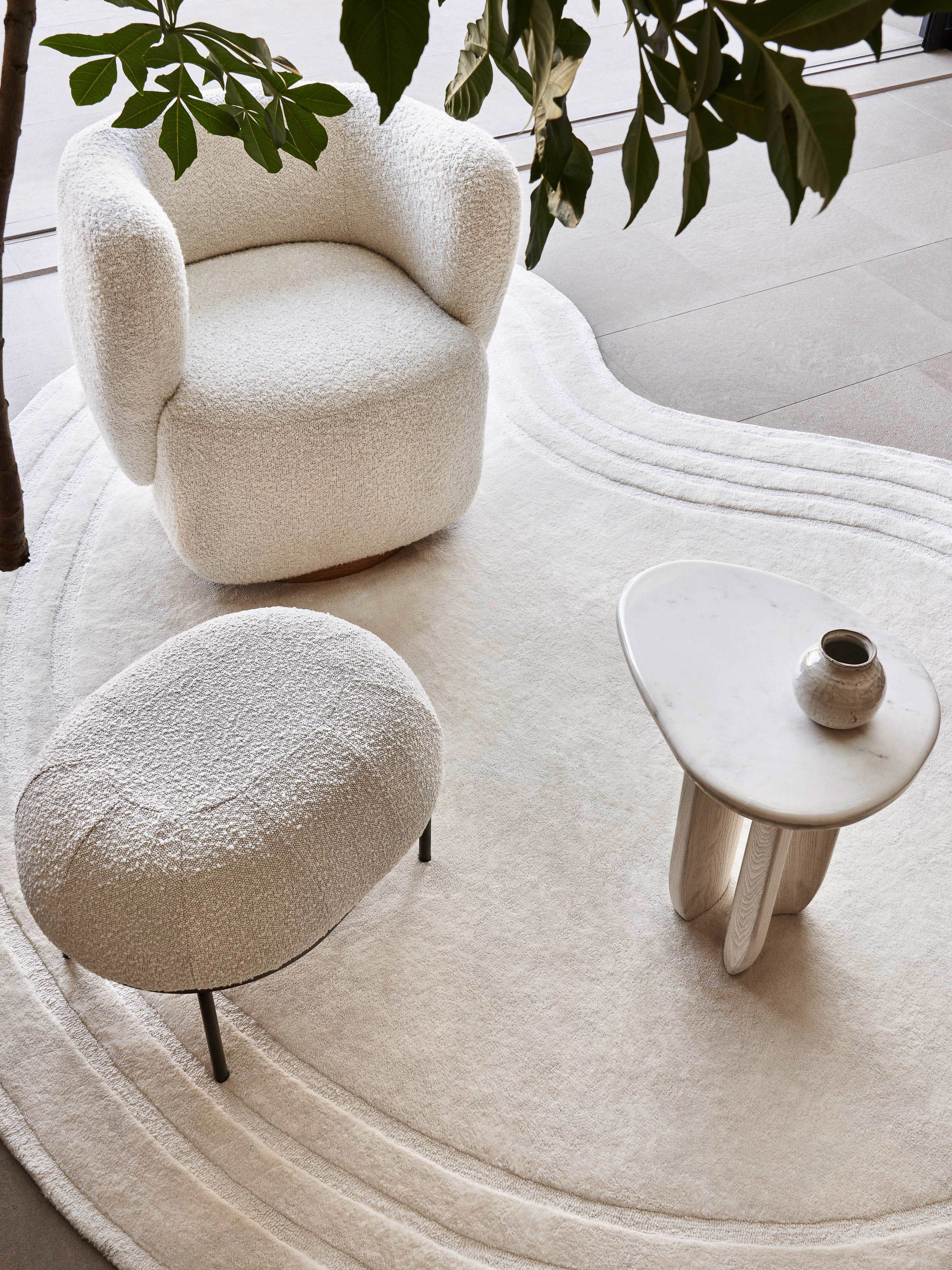 Interior design with an armchair, footstool, side table, and vase on a patterned cream rug, complemented by indoor plants for a modern, cozy feel.