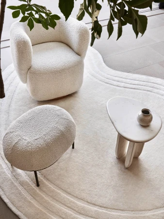 Interior design with an armchair, footstool, side table, and vase on a patterned cream rug, complemented by indoor plants for a modern, cozy feel.