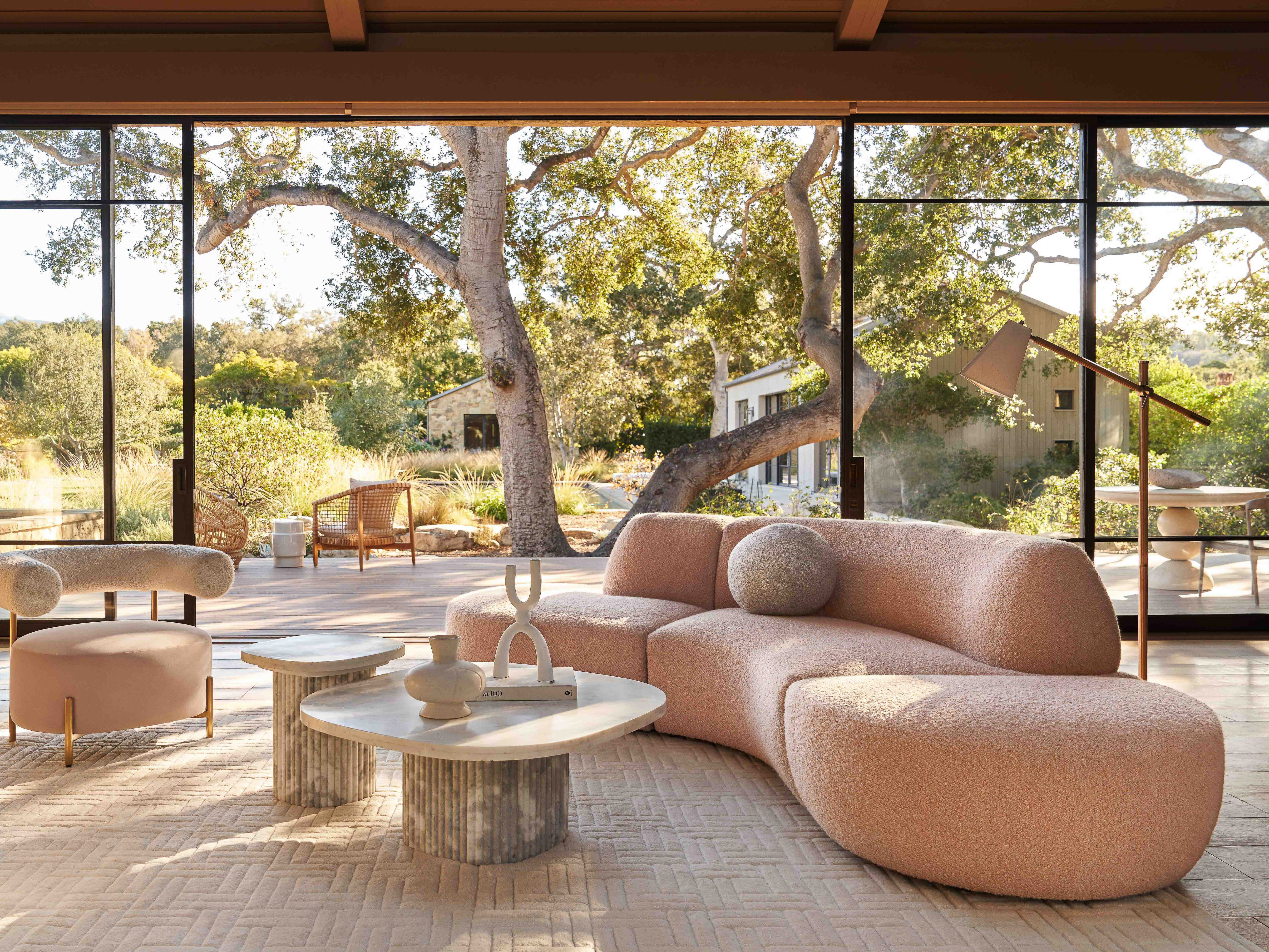 Living room interior featuring a curved pink sofa, round marble coffee table, and modern design elements, with a view of a natural landscape.