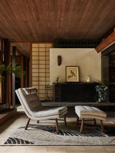Modern upholstered lounge chair and ottoman positioned on a patterned rug before a stone fireplace and shoji screen.