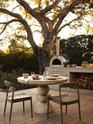 Sculptural round stone dining table set with tableware and wooden chairs on a wooden deck patio beneath a large oak tree.