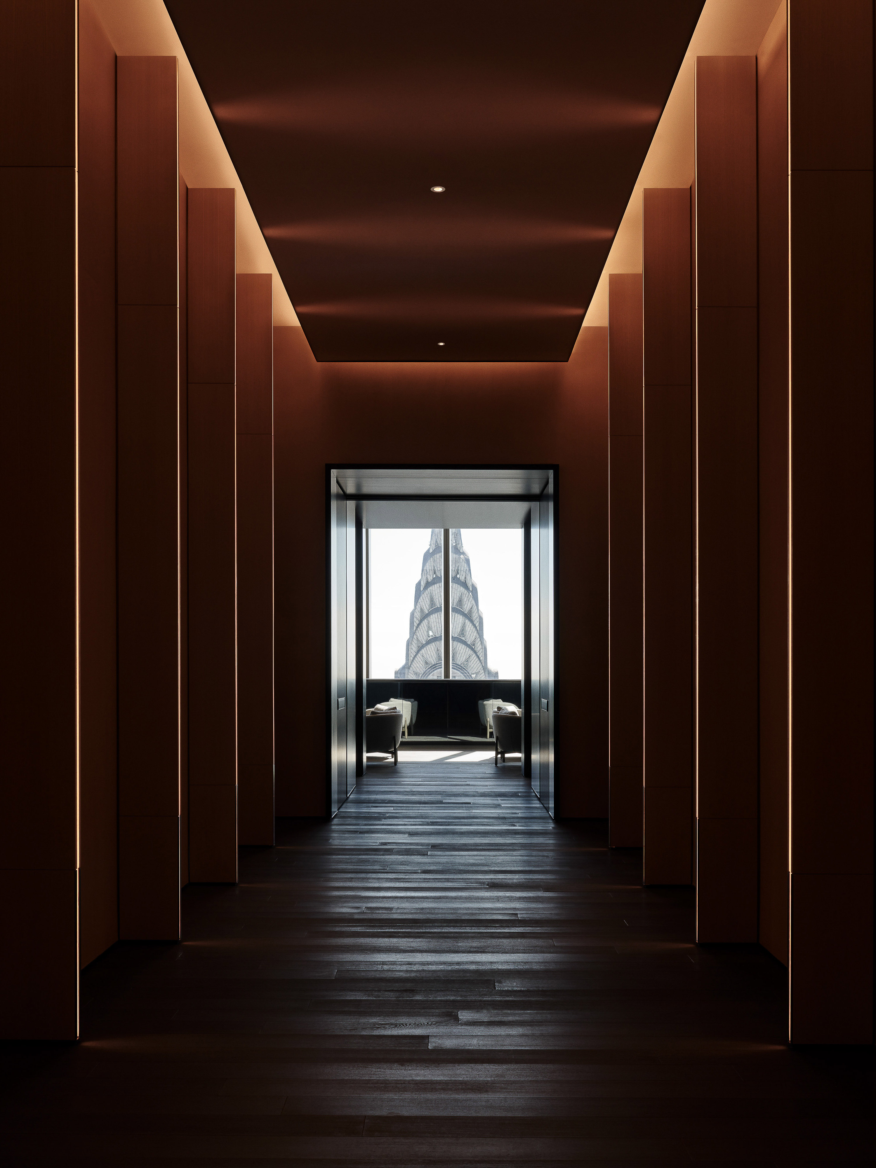Architectural view of a dark hallway with wood floors leading to a window framing the Chrysler Building, creating a sense of depth and perspective.
