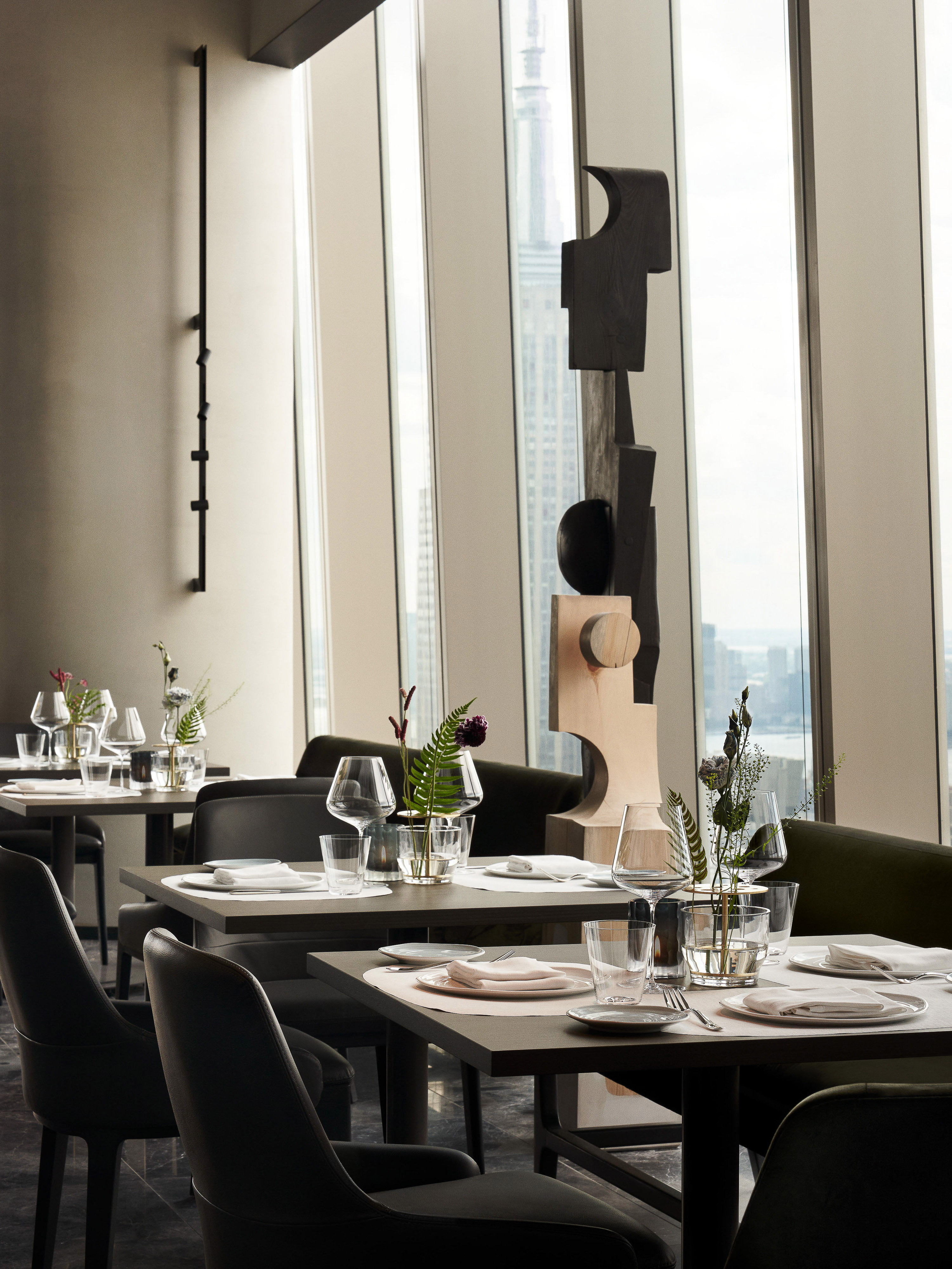 Modern restaurant interior with geometric sculpture, table settings, chairs, and natural light from large windows with a city view.