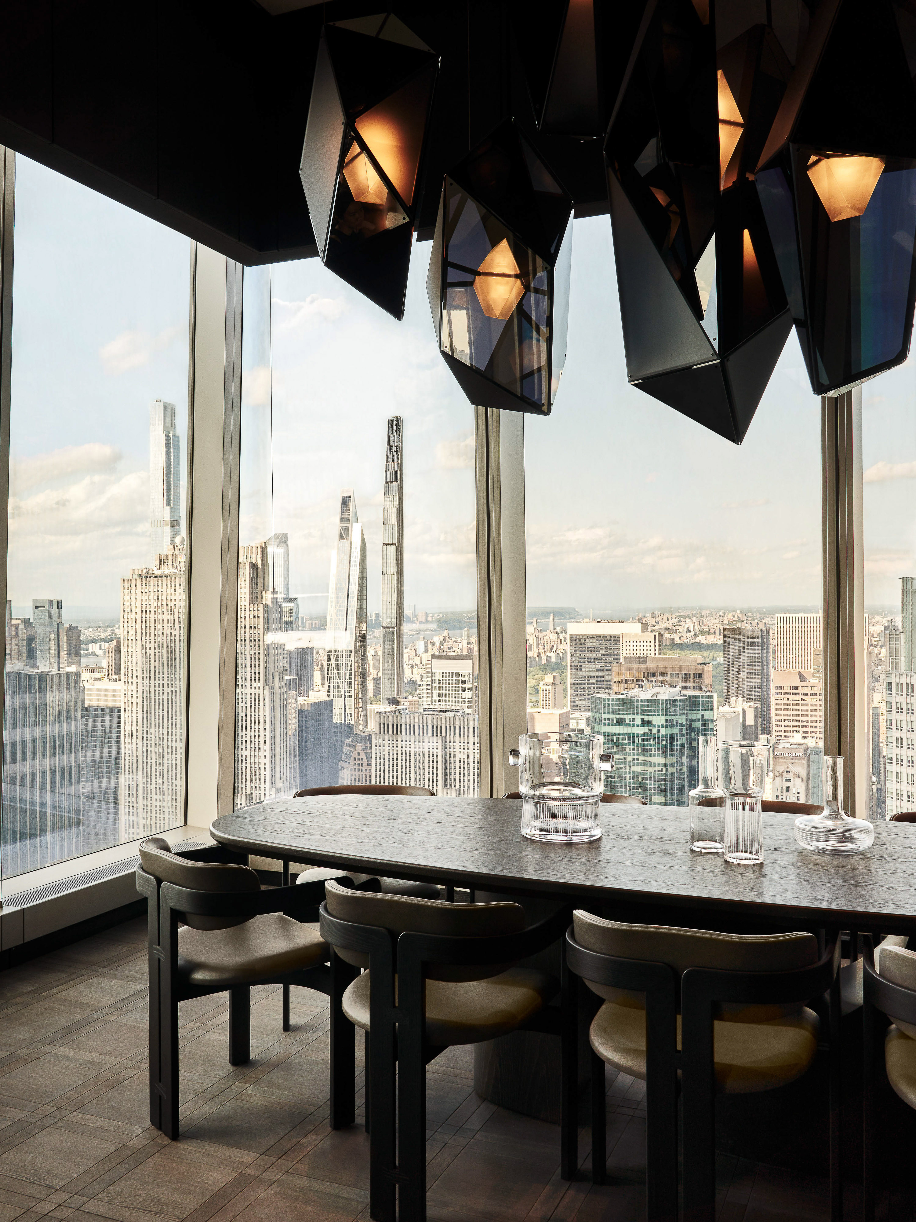 Dining room features a long oval table, geometric chandelier, and a backdrop of New York City skyline through large windows.