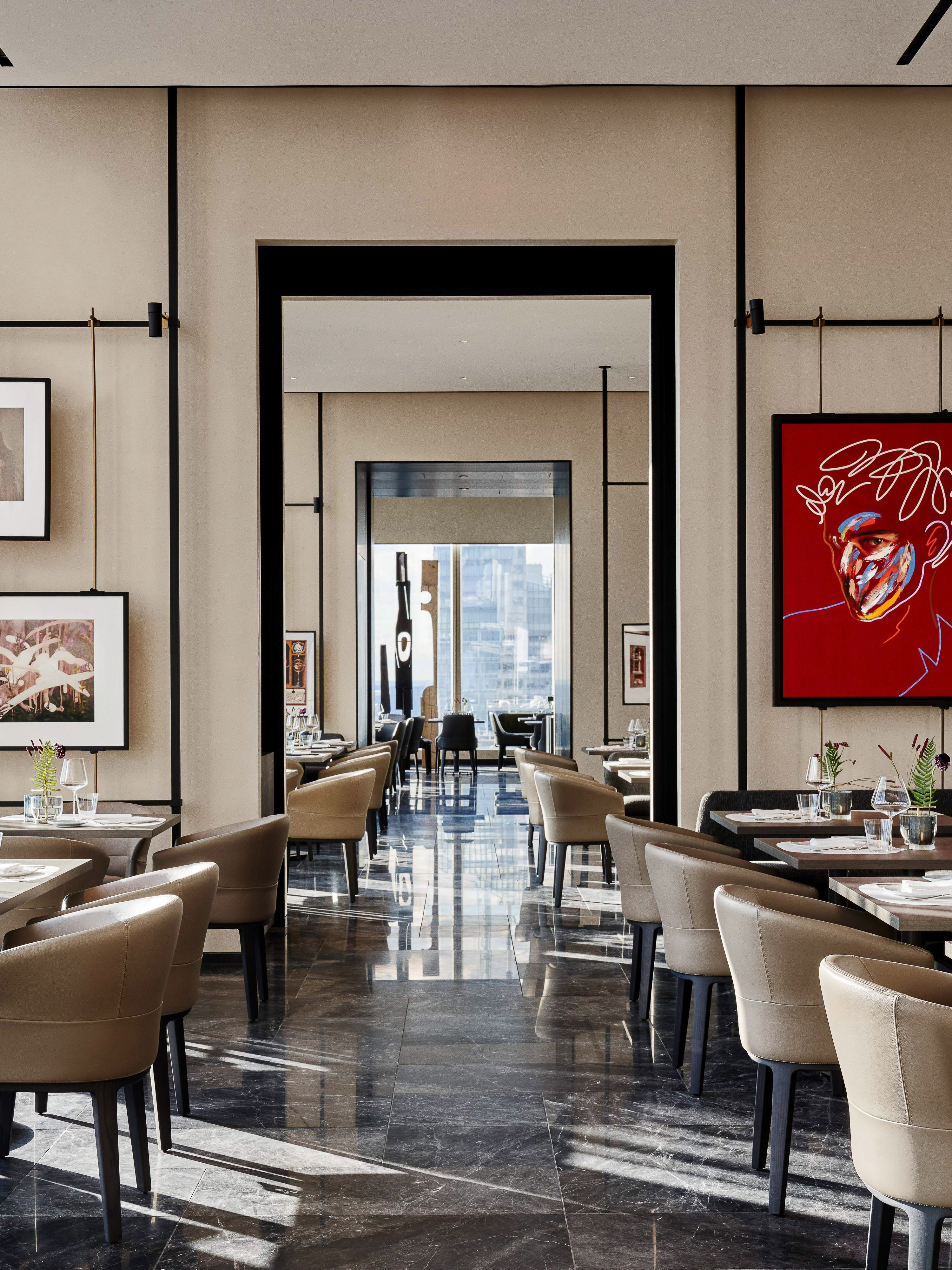 Upscale restaurant interior with marble floors, tan upholstered chairs, dining tables, artwork, and a doorway to a brighter space.