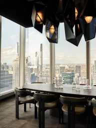 High-rise executive dining room featuring geometric pendant lights, a dark table, and expansive floor-to-ceiling city skyline views.