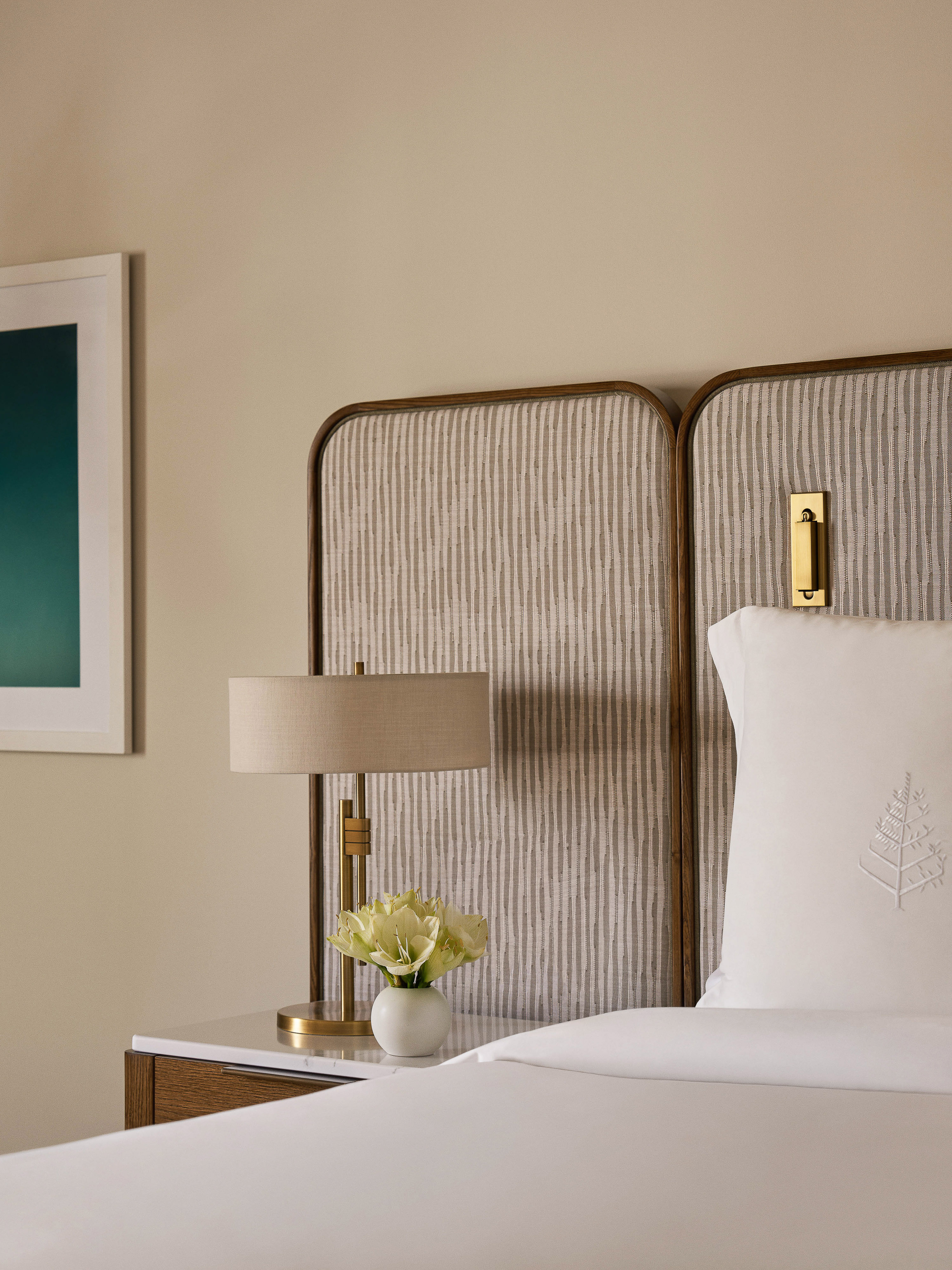 Hotel bedroom interior featuring a bed with white linens and a decorative headboard, complemented by a lamp, flowers, and framed artwork.