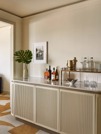 Interior view of a stylish home bar with liquor bottles, glassware, a gold cocktail shaker, and a monstera plant on a white counter.