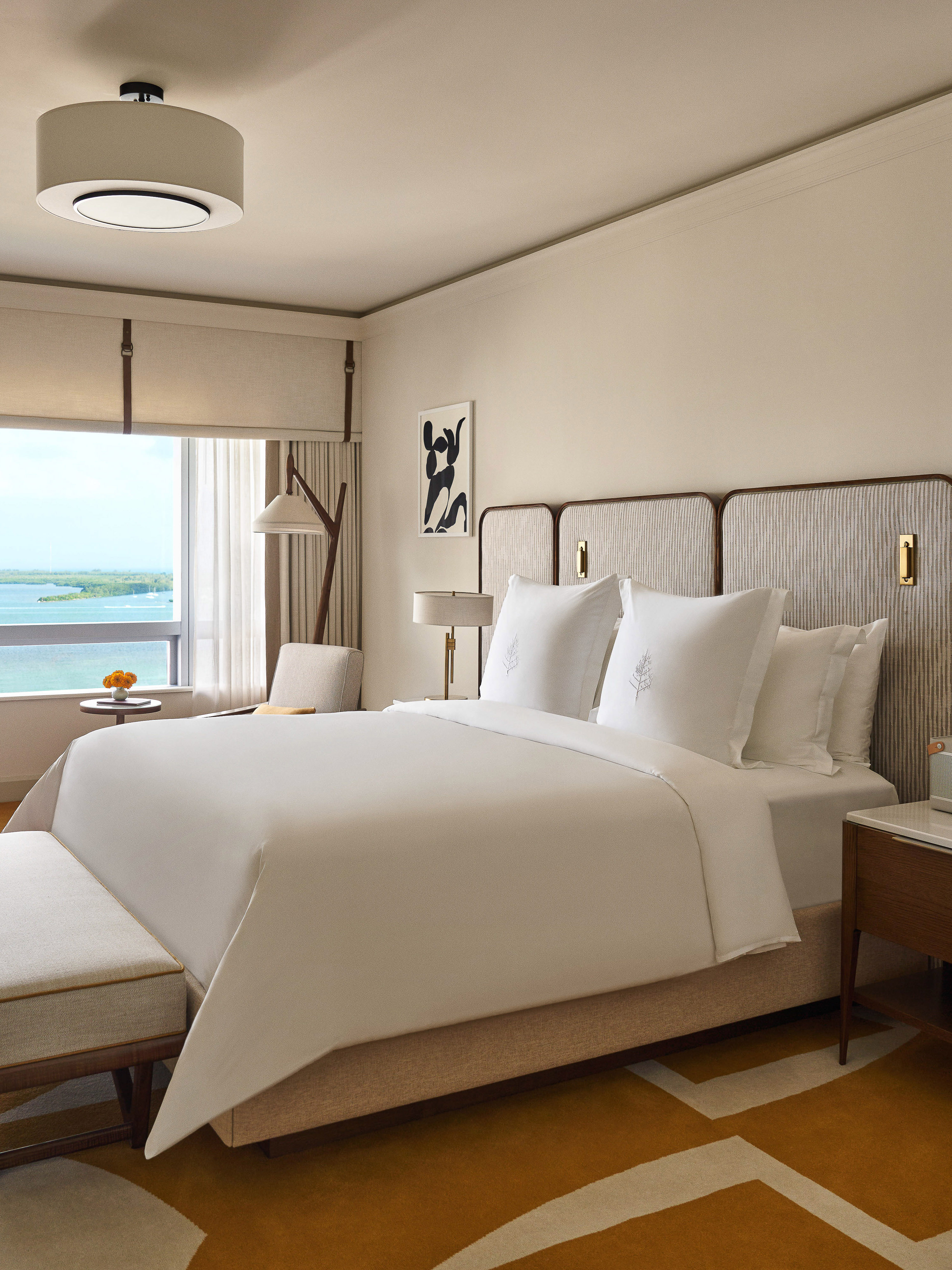 A modern hotel suite features a king-size bed with white linens, a sitting area by a window, and contemporary decor in neutral tones.
