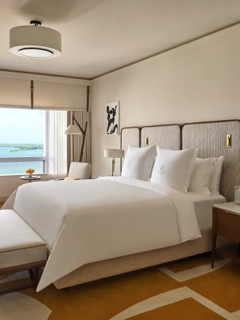 A modern hotel suite features a king-size bed with white linens, a sitting area by a window, and contemporary decor in neutral tones.
