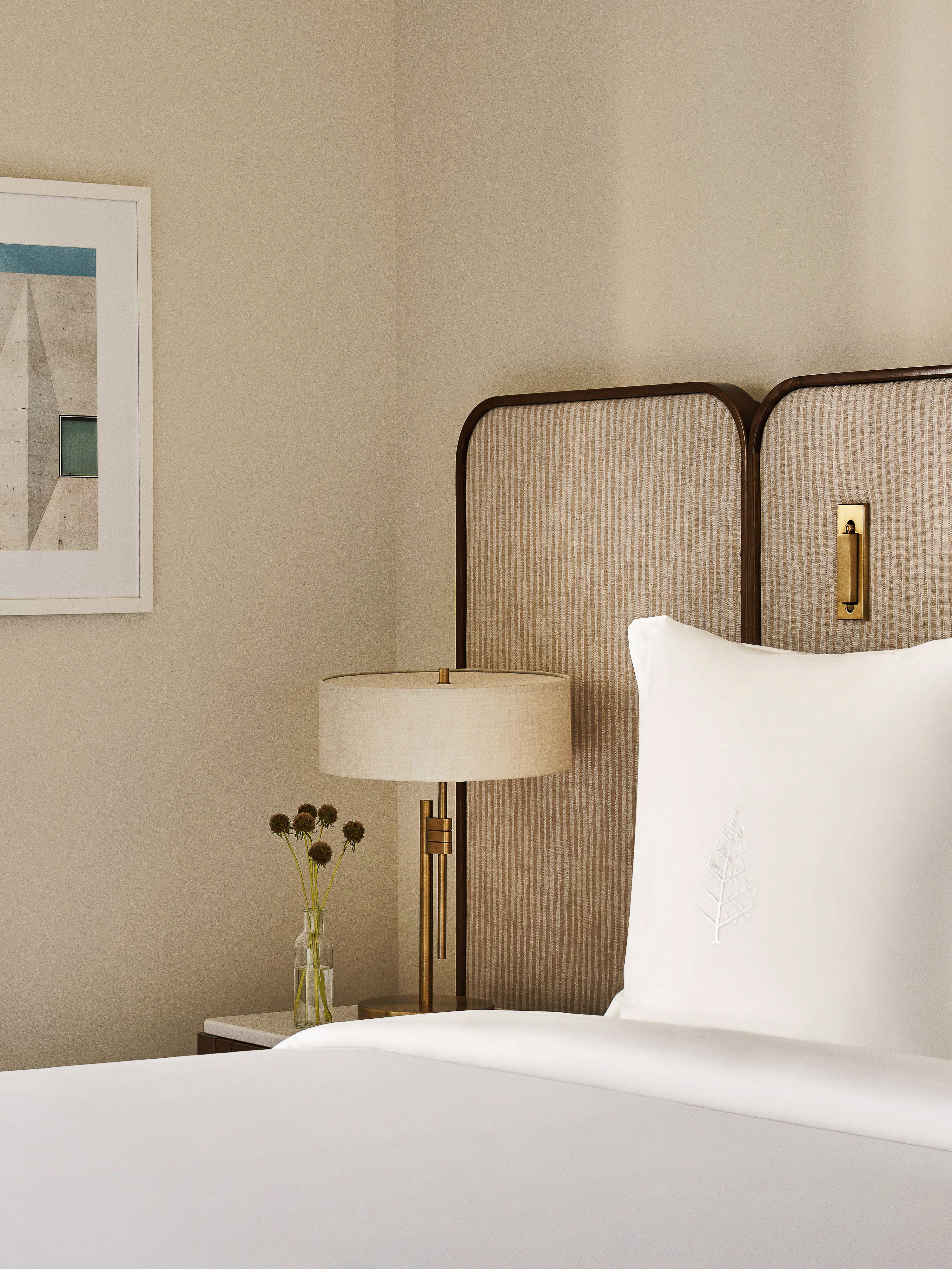 Bedroom interior featuring a bed with white linens, a textured headboard, bedside table with a lamp, and framed wall art in a neutral color palette.