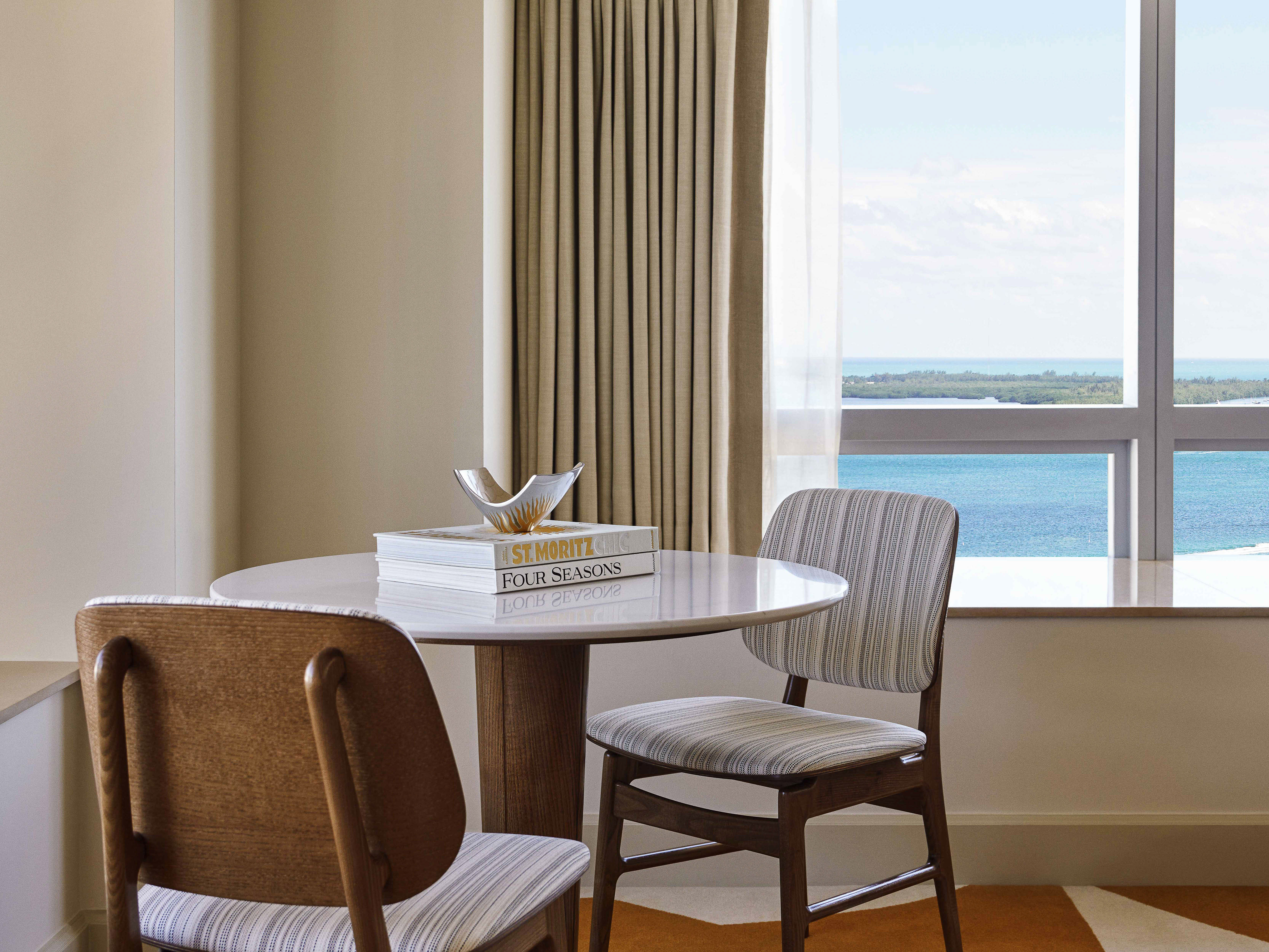 Interior shot of a seating area with a round marble table, wooden chairs, and a view of the ocean from the window in the background.