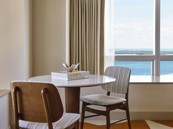 Interior shot of a seating area with a round marble table, wooden chairs, and a view of the ocean from the window in the background.