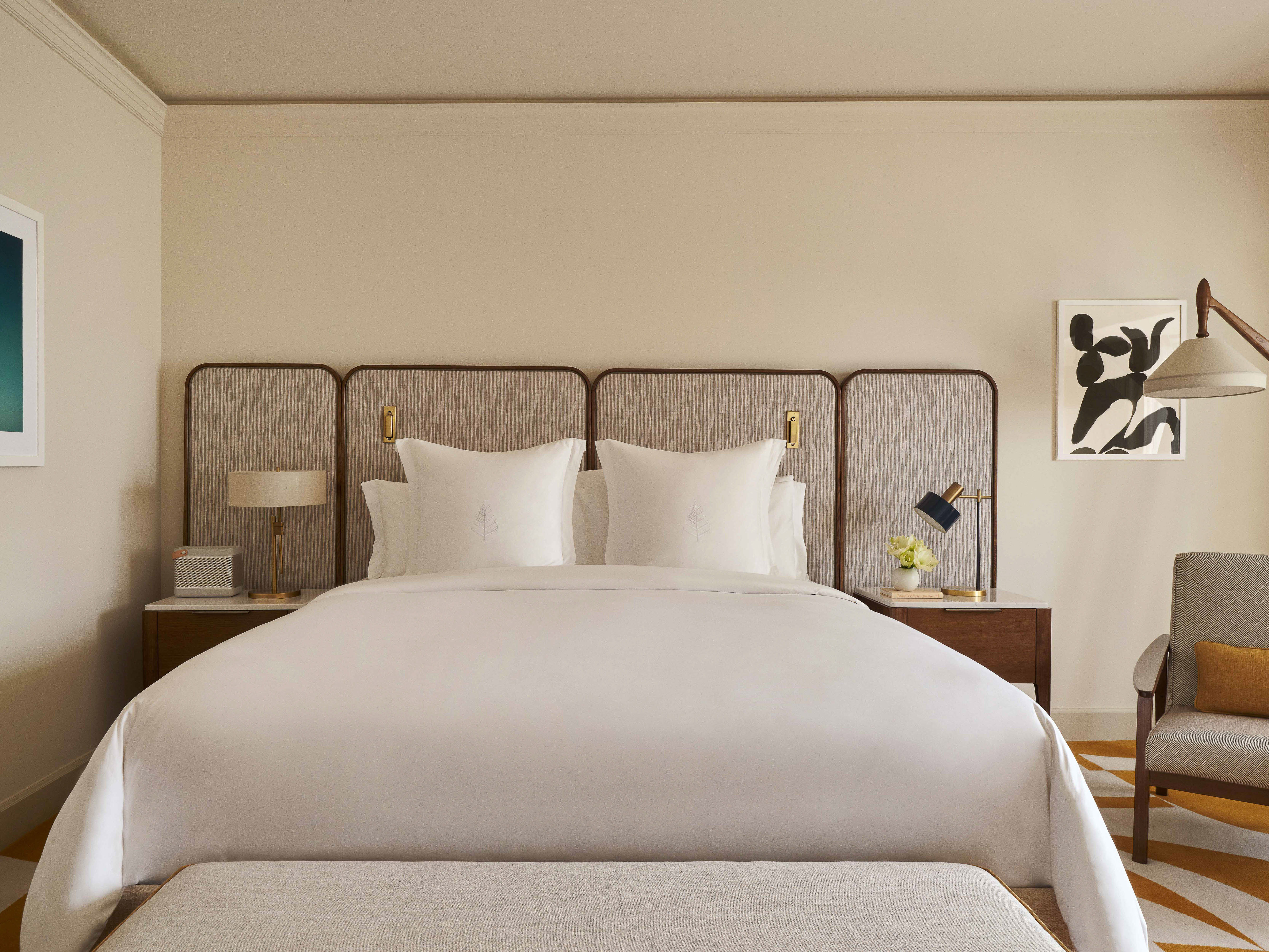 Bedroom interior featuring a made-up bed with white linens, a textured headboard, a patterned carpet, and neutral-toned walls with framed artwork.