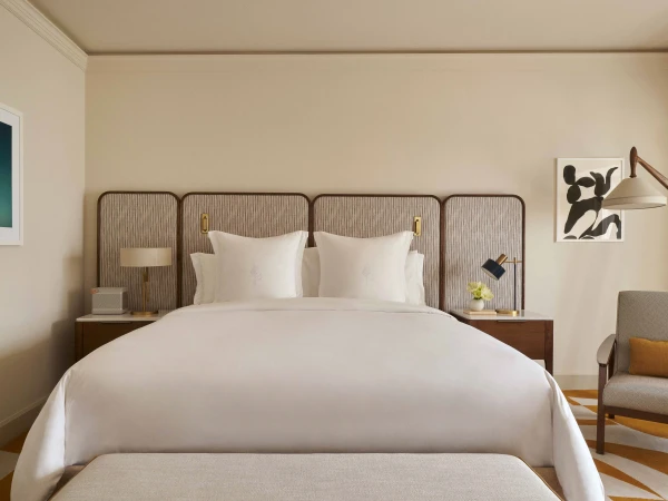 Bedroom interior featuring a made-up bed with white linens, a textured headboard, a patterned carpet, and neutral-toned walls with framed artwork.