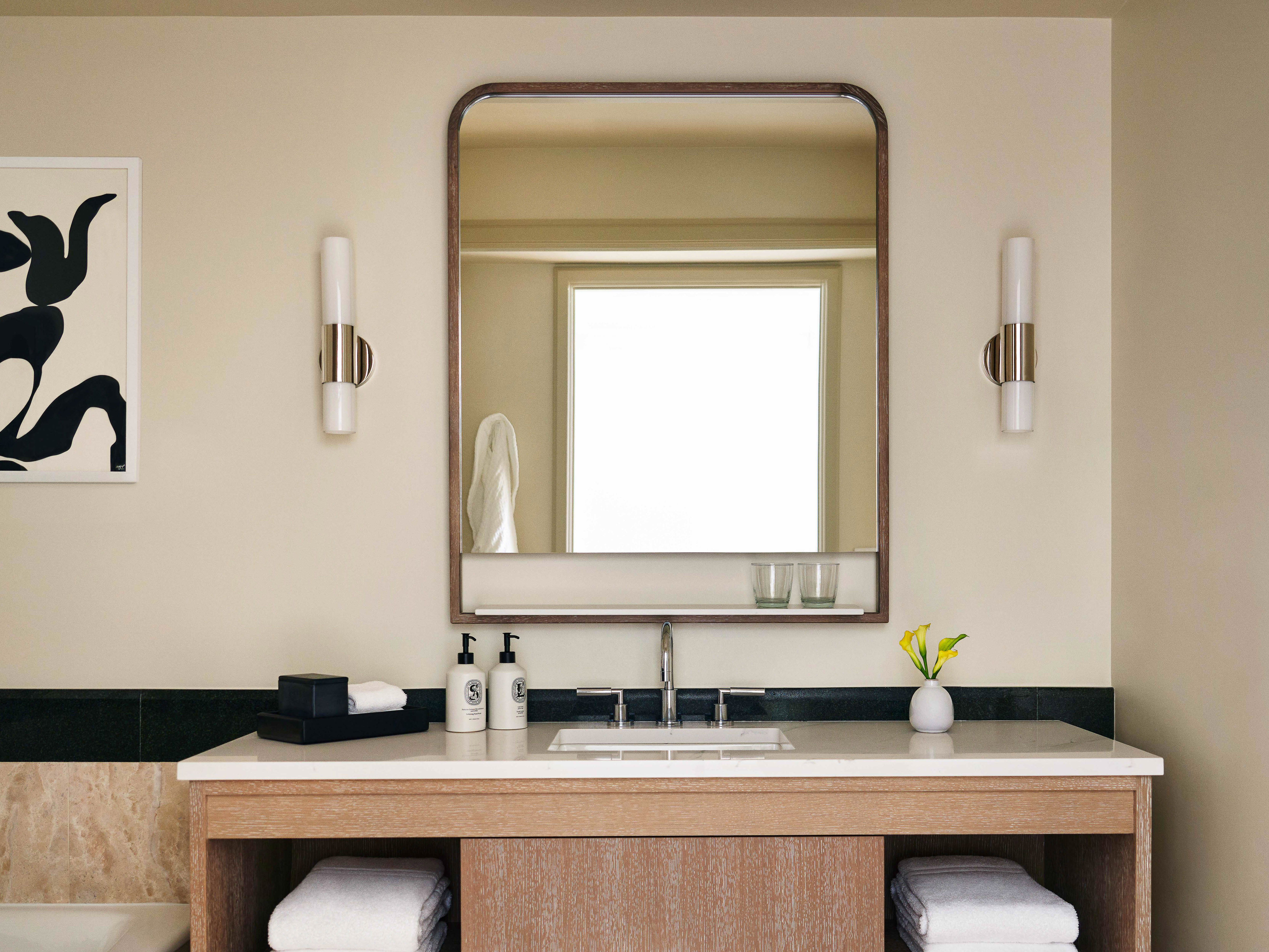 Bathroom interior with a wooden vanity, white countertop, mirror, wall-mounted sconces, and a framed abstract art piece on the wall.