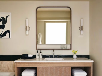 Modern hotel bathroom vanity with light wood cabinetry, white stone countertop, rectangular mirror, and twin wall sconces.