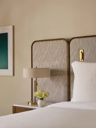 Four Seasons hotel bed with white embroidered pillow, textured headboard, brass nightstand lamp, and floral vase.