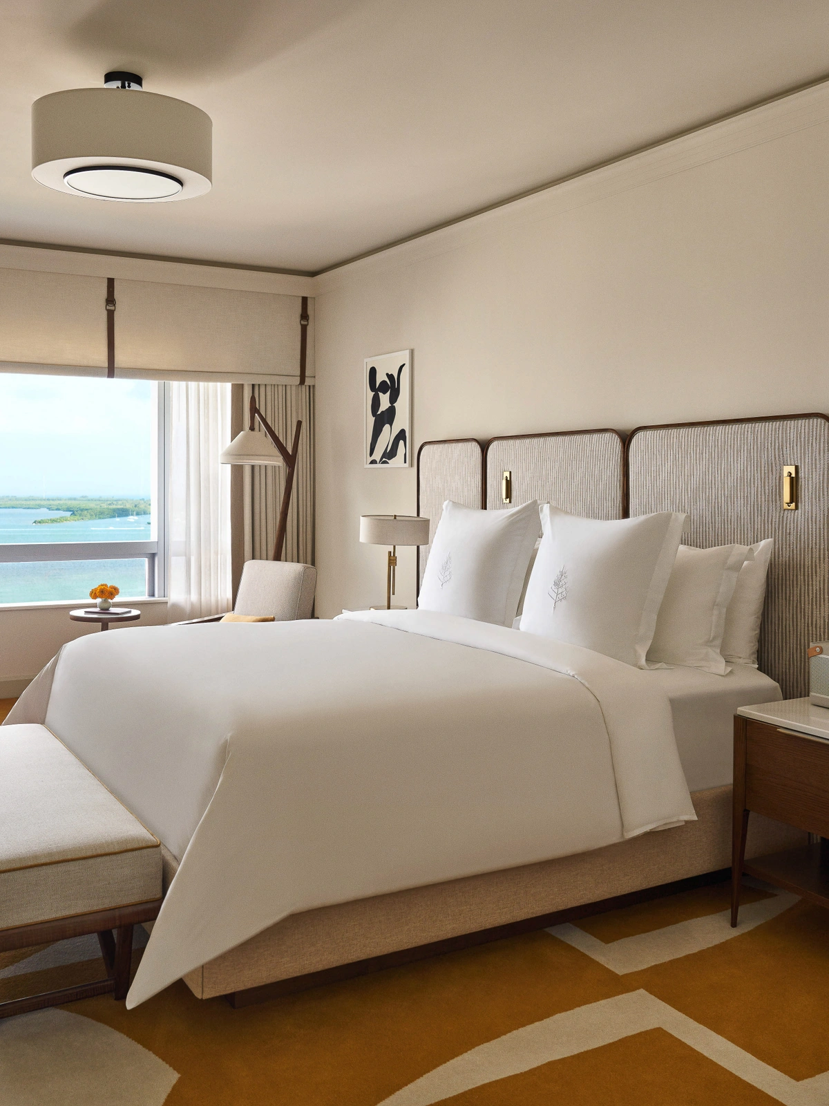A dressed king bed with crisp white linens and a textured upholstered headboard beside an ocean view window in a luxury hotel room.