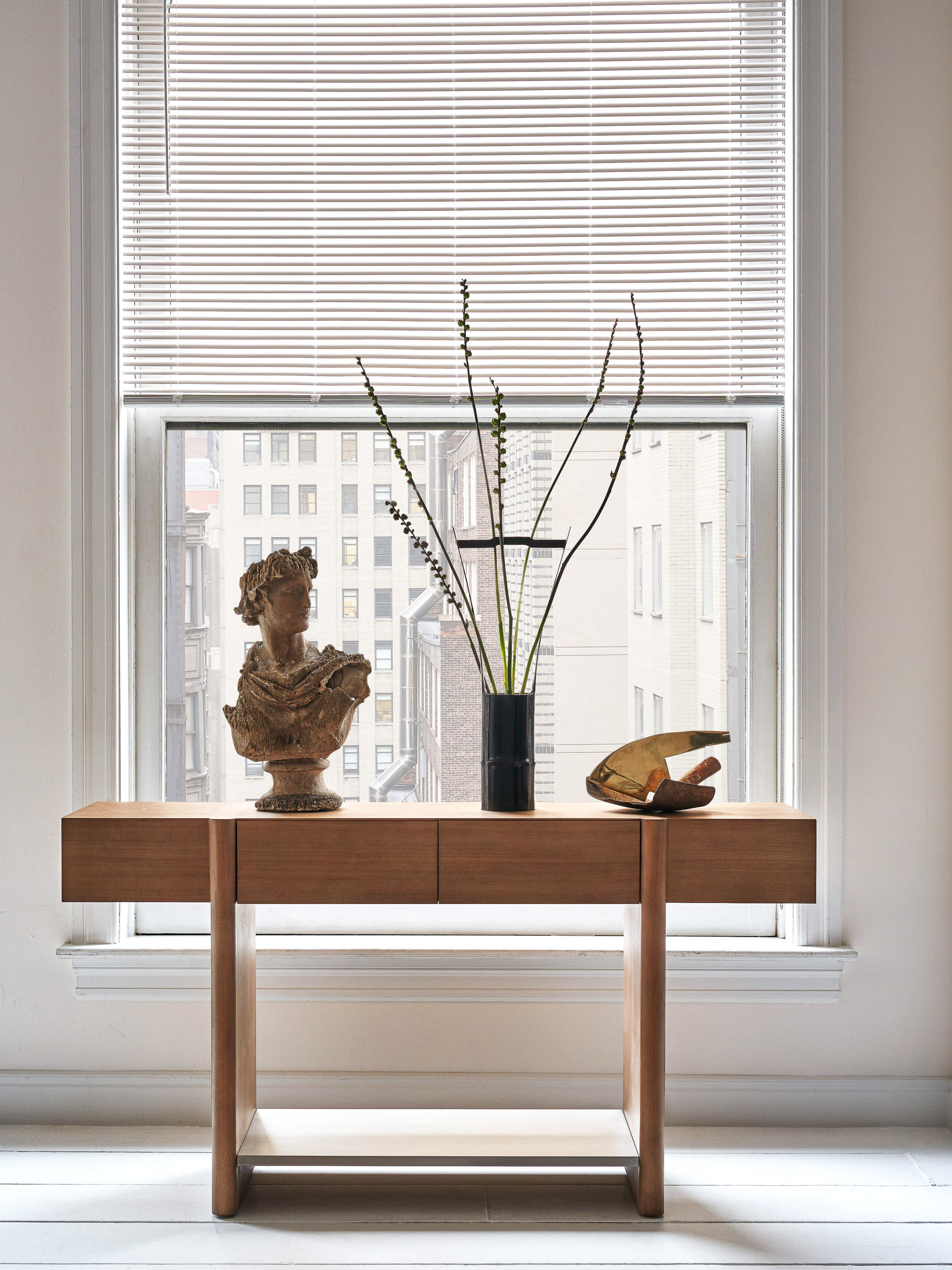 Interior with a wooden console table, a bust sculpture, decorative branches in a vase, and an amber glass bowl in front of a window.