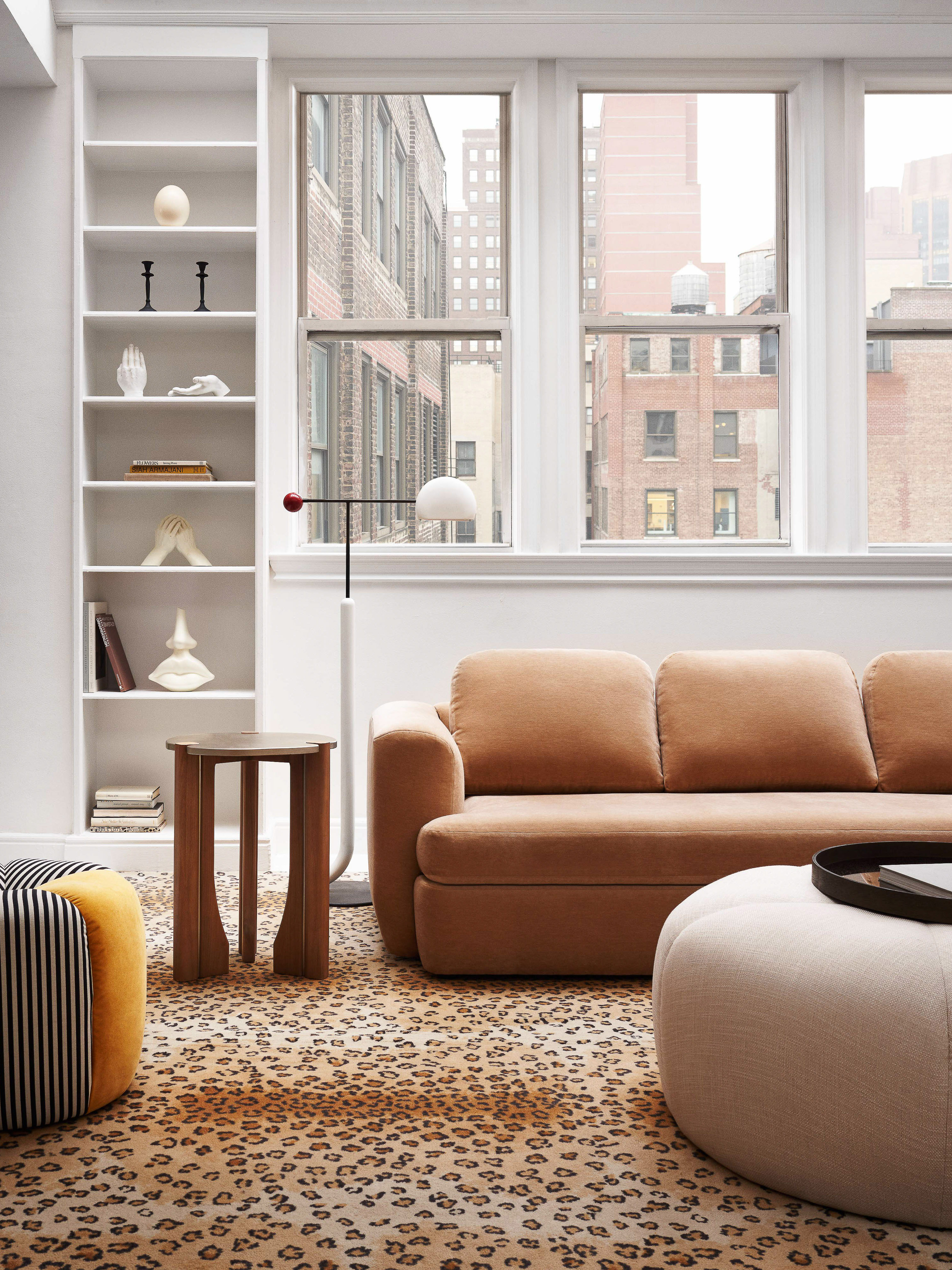 Living room interior featuring a caramel-colored sofa, side table, patterned rug, white shelving, and a view of buildings through a window.