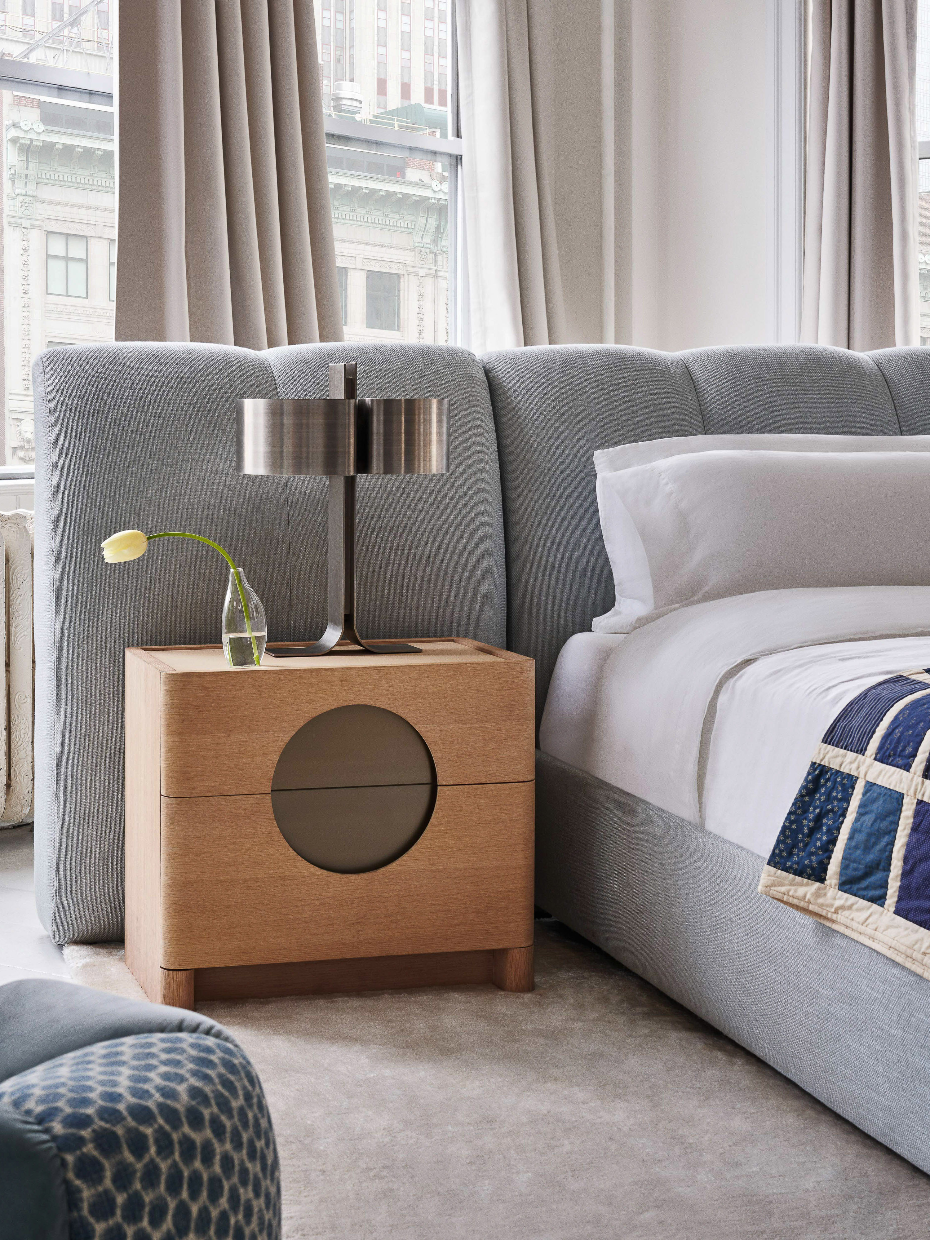 Modern bedroom interior featuring a light blue upholstered bed with a patterned quilt, wooden nightstand with a lamp, and light curtains.
