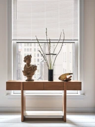 Wooden console table with two drawers placed in front of a large window with blinds, styled with a classical bust and vase.