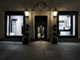 Exterior view of a luxury stone storefront at night featuring illuminated Saint Laurent window displays, glowing lanterns, and potted topiaries.