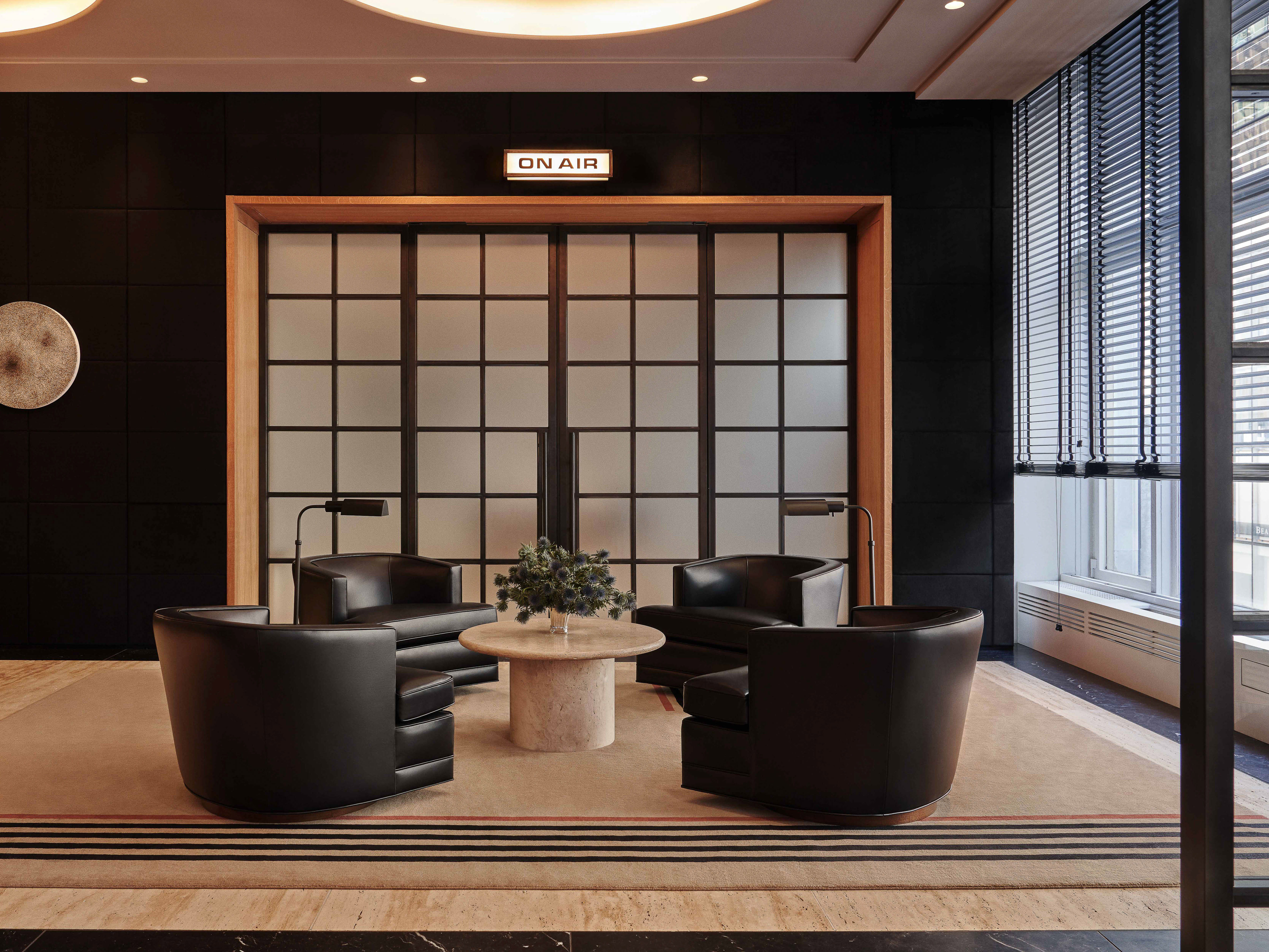Four black leather armchairs arranged around a round stone coffee table in a studio lounge beneath an illuminated On Air sign.