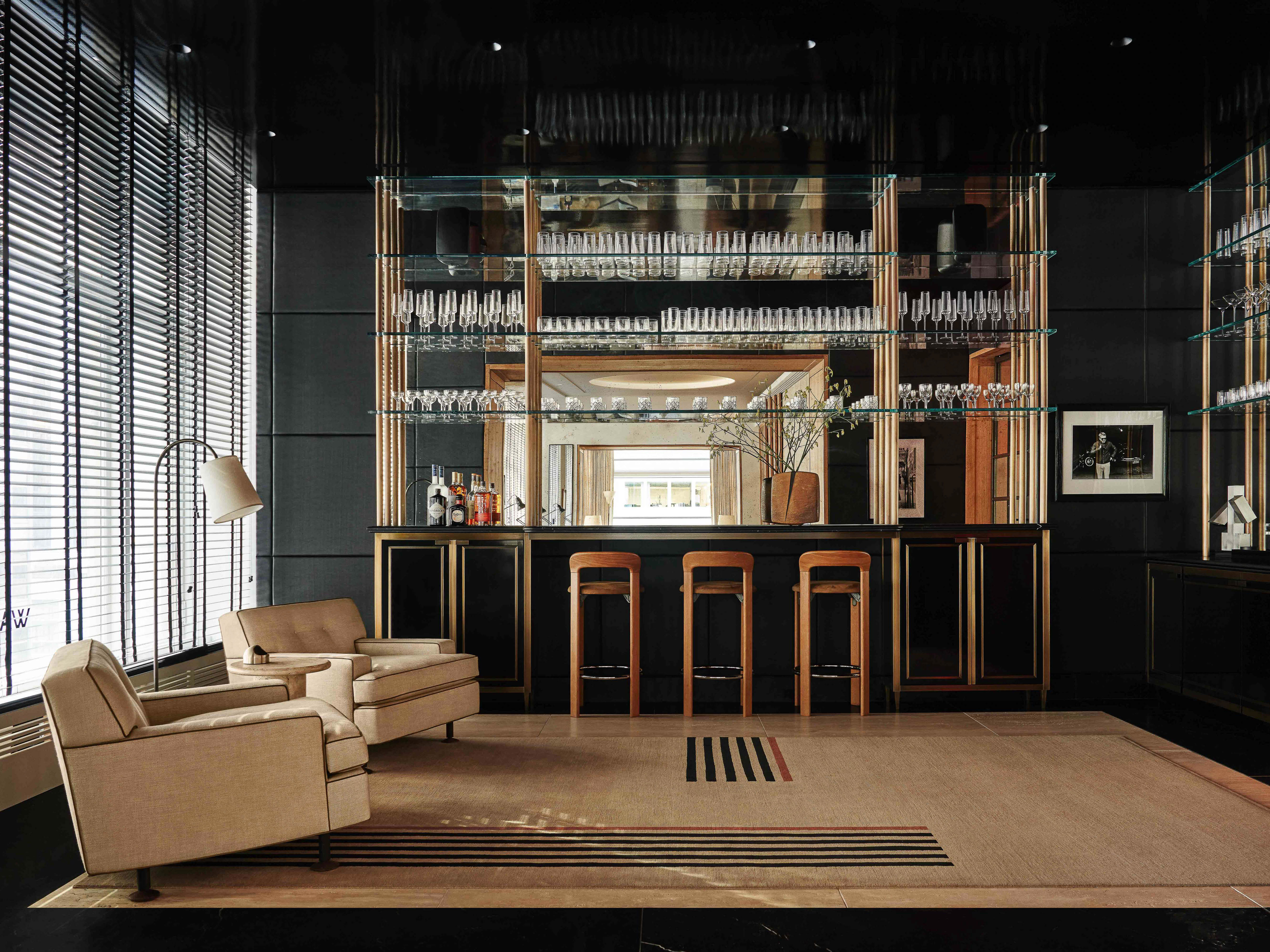 Interior shot of a home bar area with two armchairs, a bar with barstools, glass shelving, and window blinds, bathed in warm light.