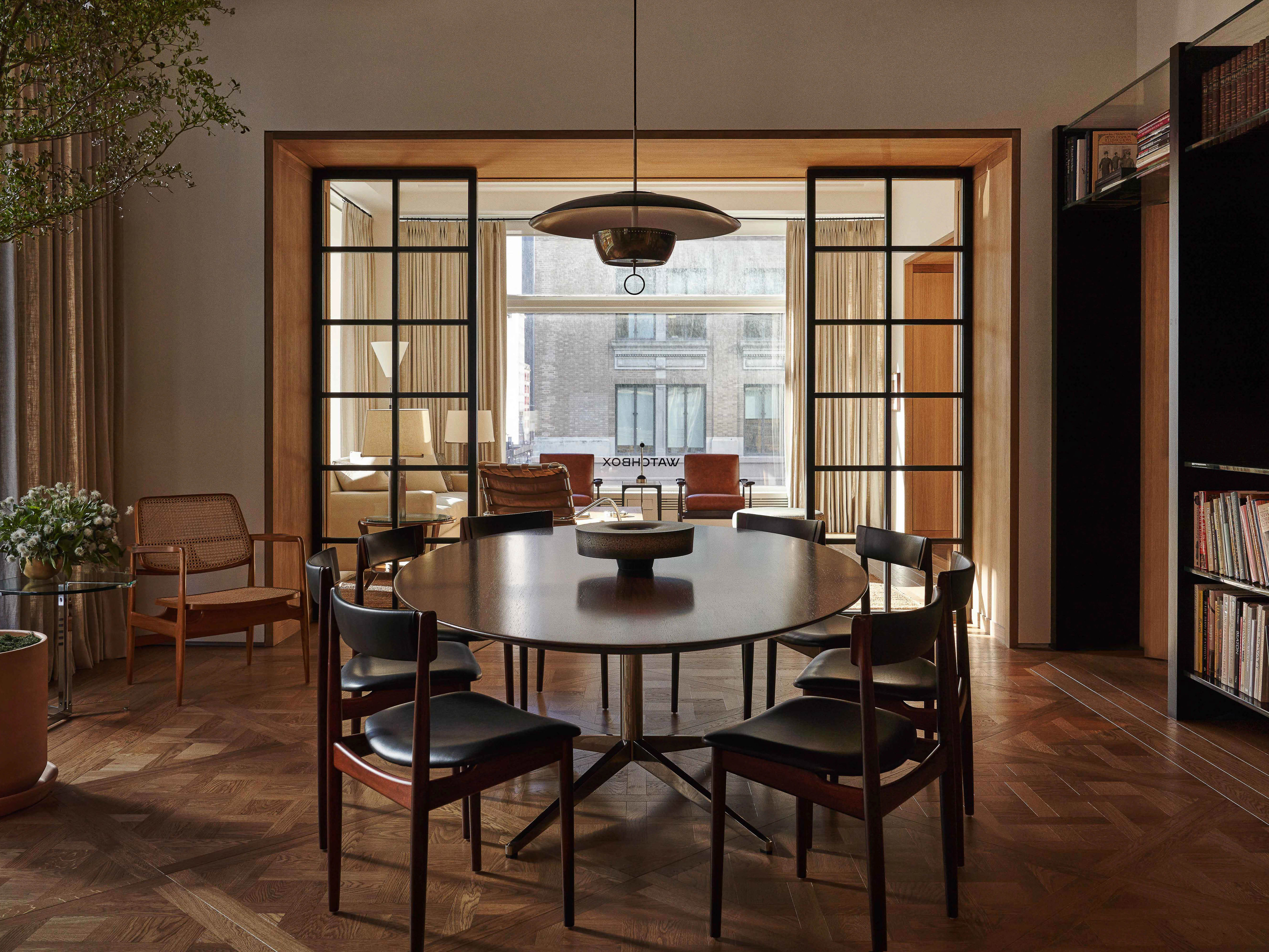Interior design of a modern dining room featuring a wood table, chairs, herringbone floor, black shelving, and natural light from windows.