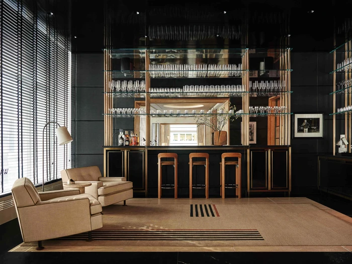 A luxury bar lounge interior featuring a sleek black counter with three wooden barstools, glassware shelving, and two beige armchairs.