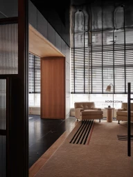 Modern luxury showroom lounge featuring beige armchairs, a geometric rug, wood doorway framing, and horizontal window blinds.