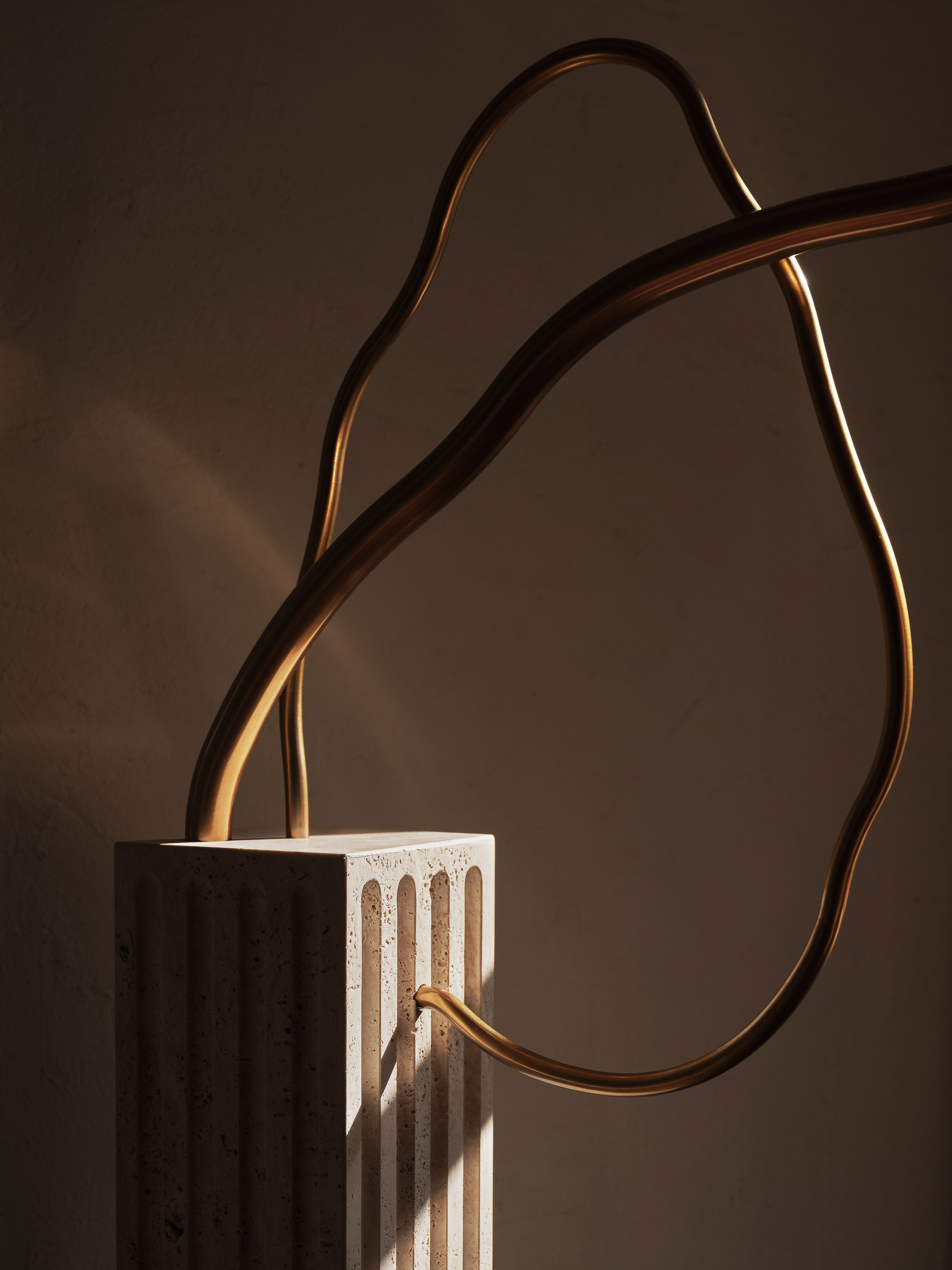 Abstract sculpture featuring a fluted column base with a curved, golden metal rod structure in a warm, minimalist setting.
