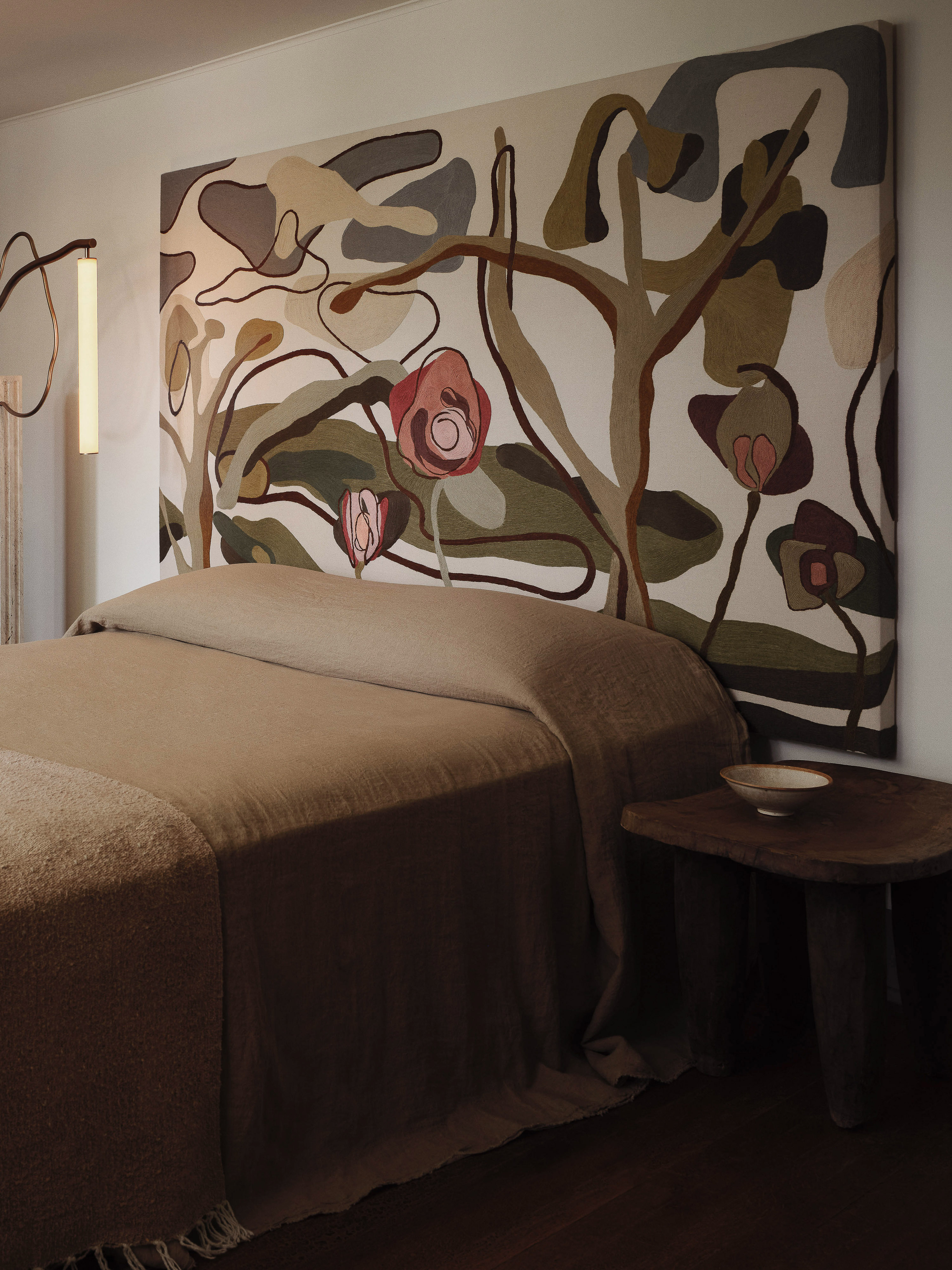 Bedroom features a tan bed with a neutral-toned, abstract floral artwork headboard and a wooden side table with a small bowl.