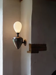 Sculptural bronze wall sconce with a glowing white oval glass shade mounted on an off-white plaster wall with an exposed wood beam.