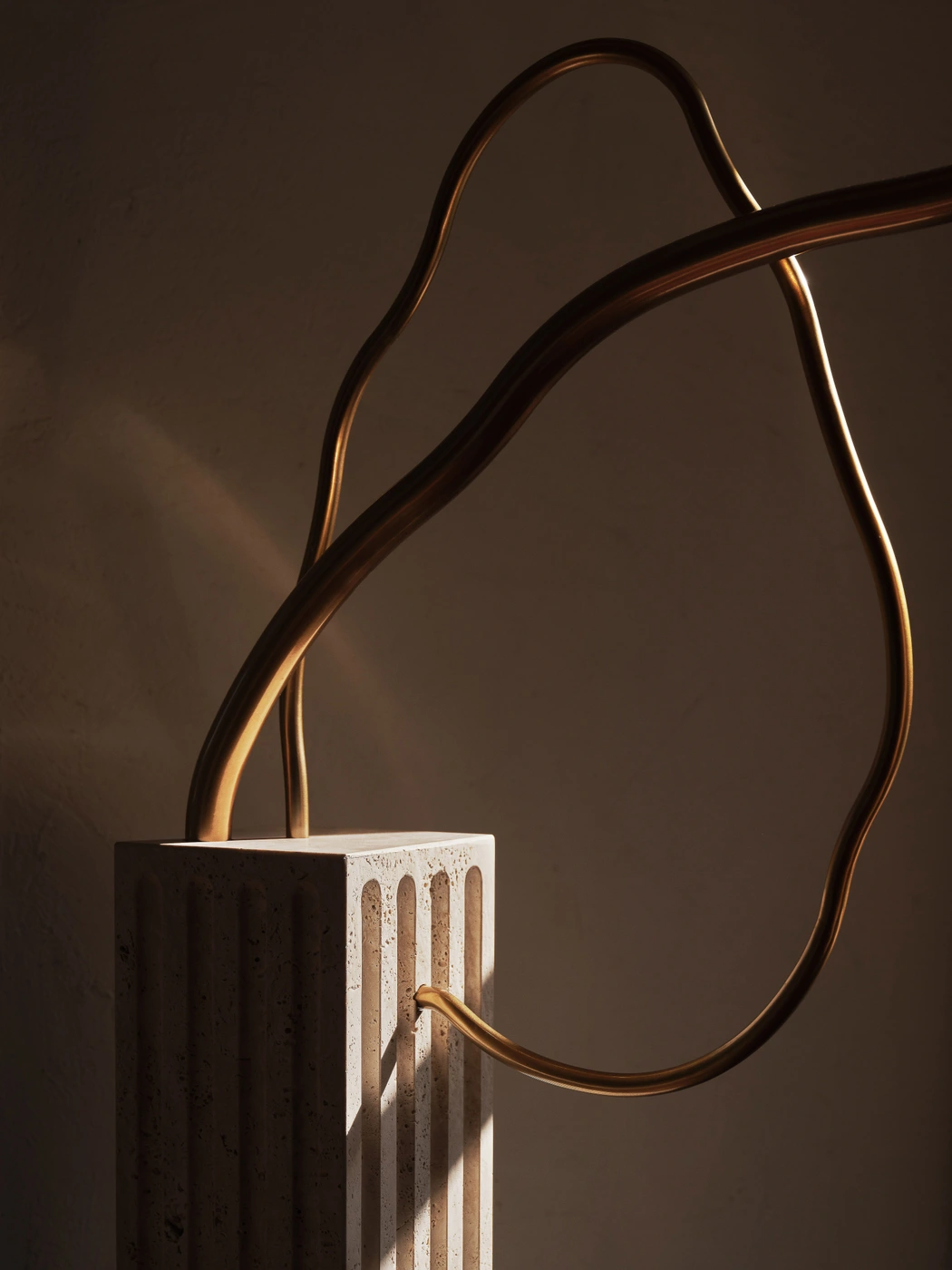Sculptural fluted stone plinth with curved bronze metal tubes looping upward against a warm neutral wall with soft shadows.