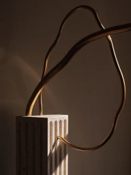 Sculptural fluted stone plinth with curved bronze metal tubes looping upward against a warm neutral wall with soft shadows.