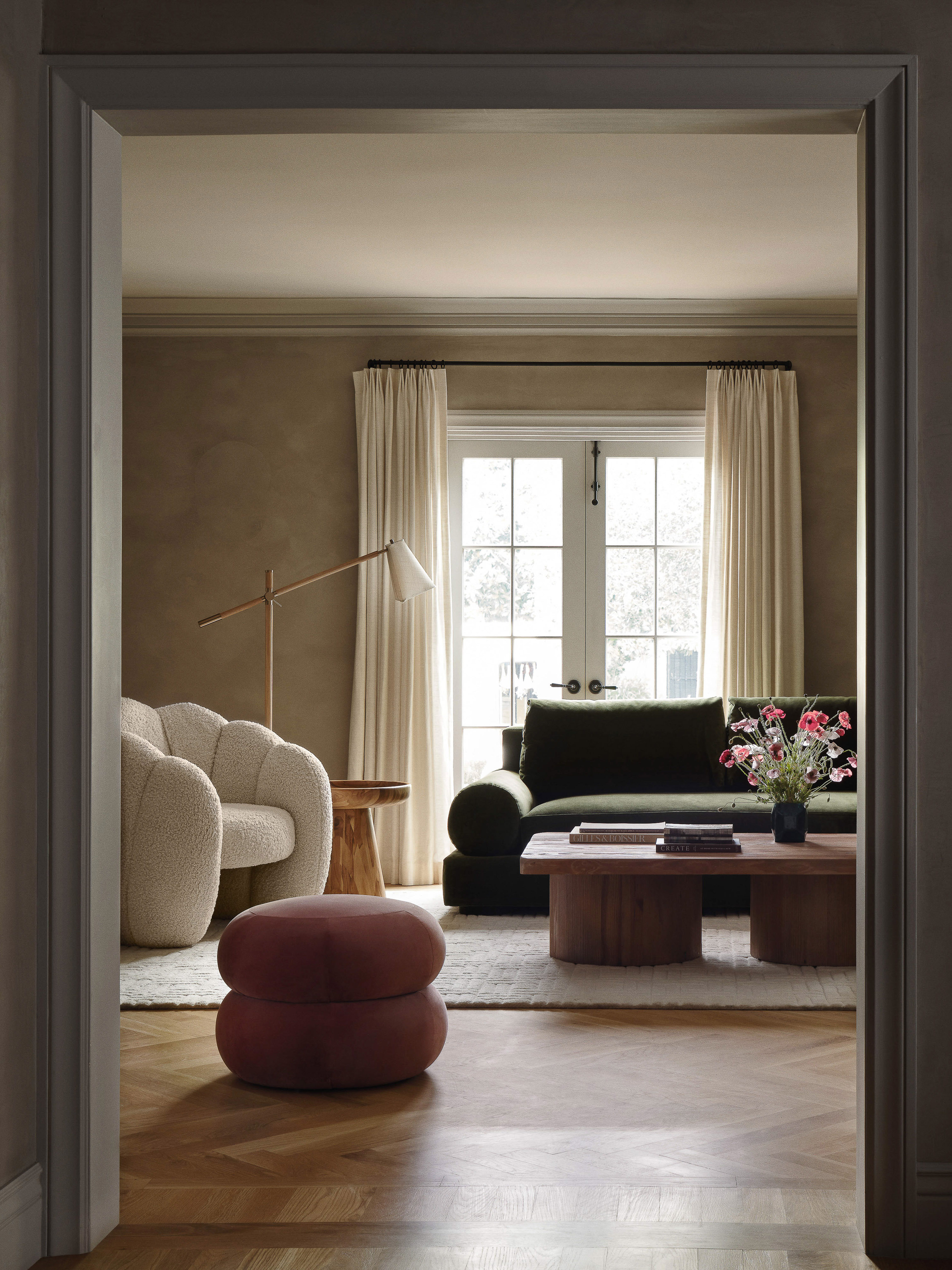 View through an open doorway into a styled living room with a green velvet sofa, cream boucle chair, and round pink pouf.