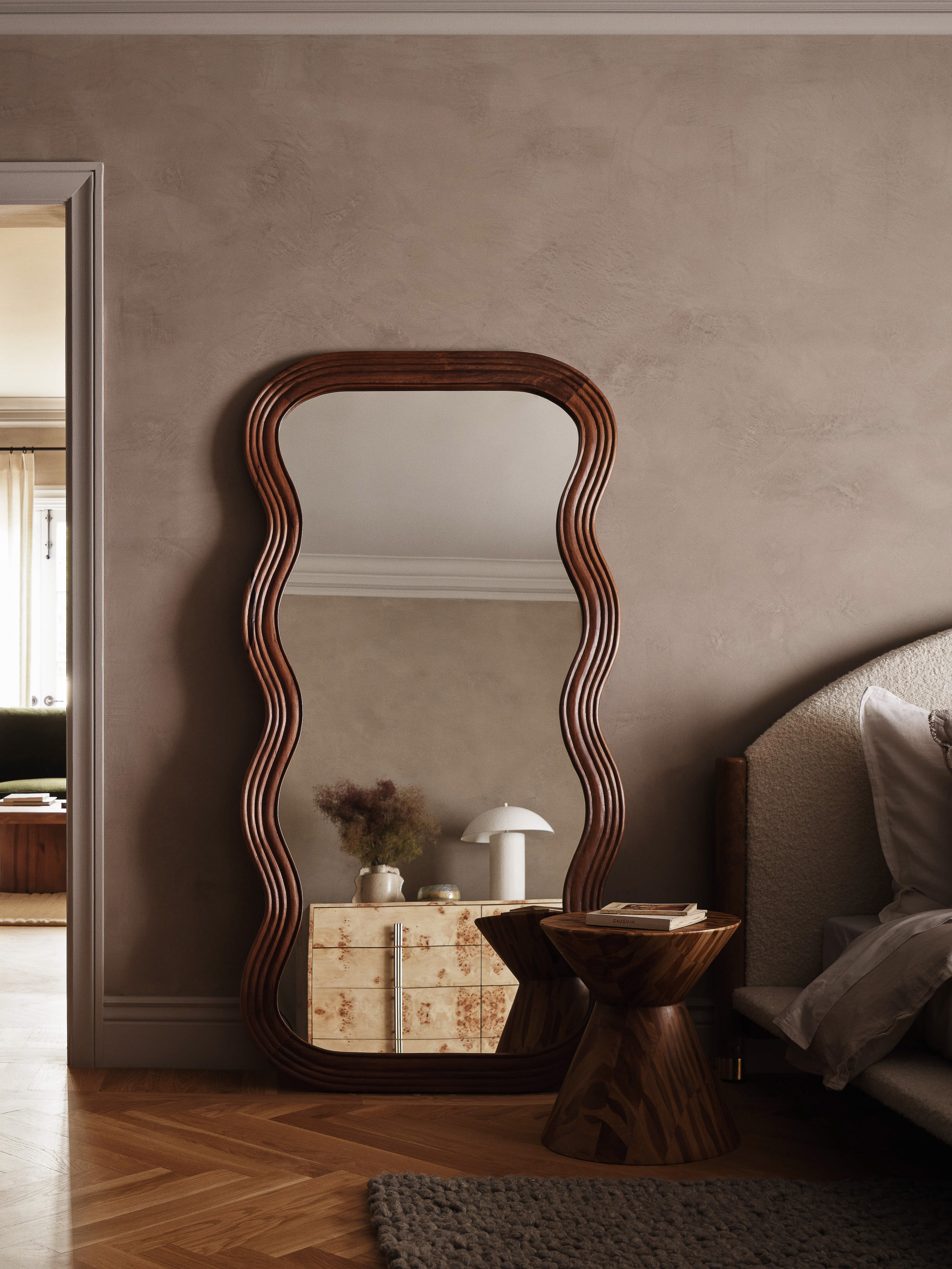 Interior view featuring a wavy framed mirror, a wooden side table with books, a patterned dresser, and parquet flooring in a neutral palette.
