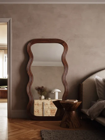Interior view featuring a wavy framed mirror, a wooden side table with books, a patterned dresser, and parquet flooring in a neutral palette.