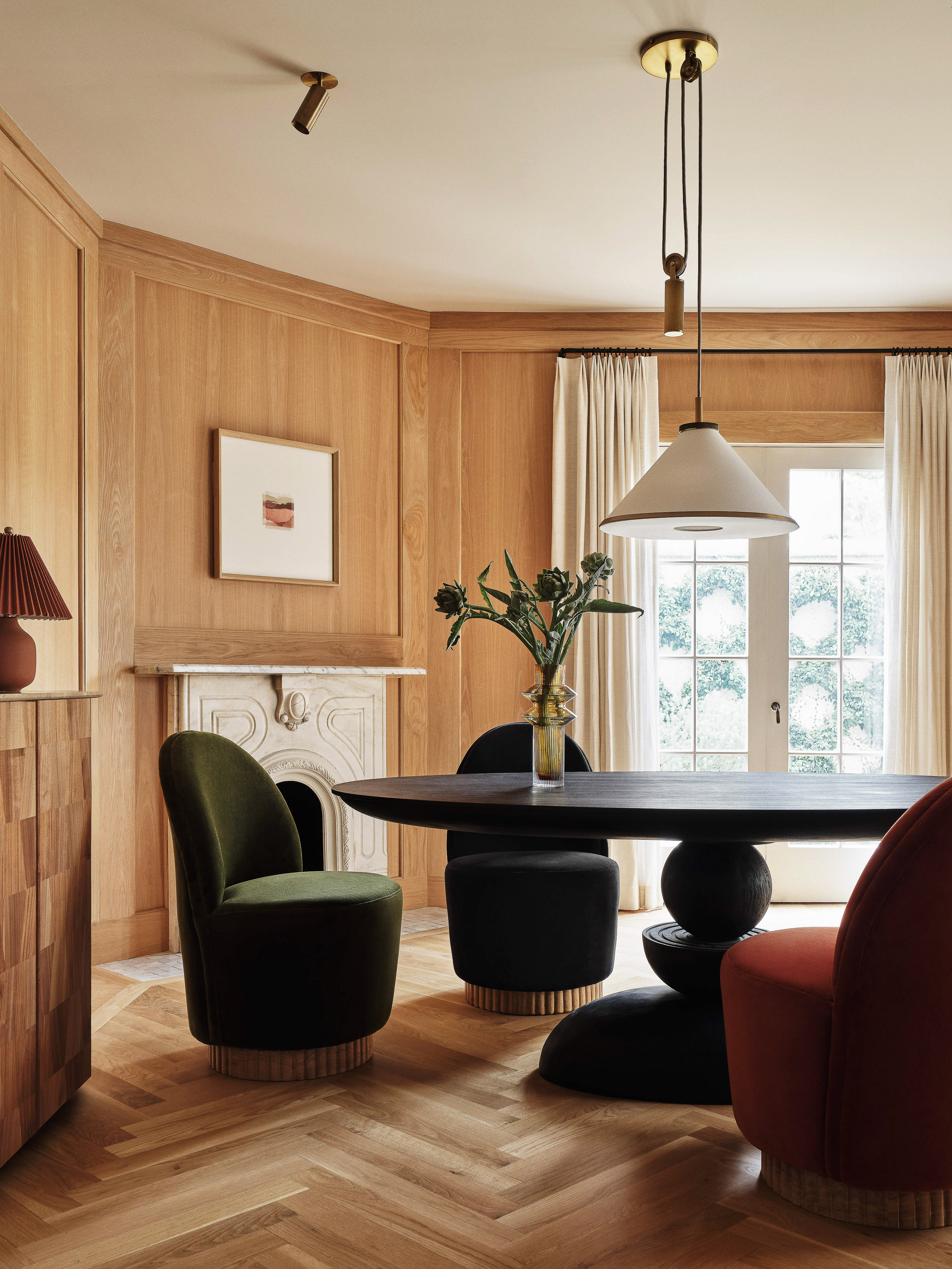 Interior of a dining room with wood paneled walls, a dark wood dining table, olive green chairs, and white curtains. 