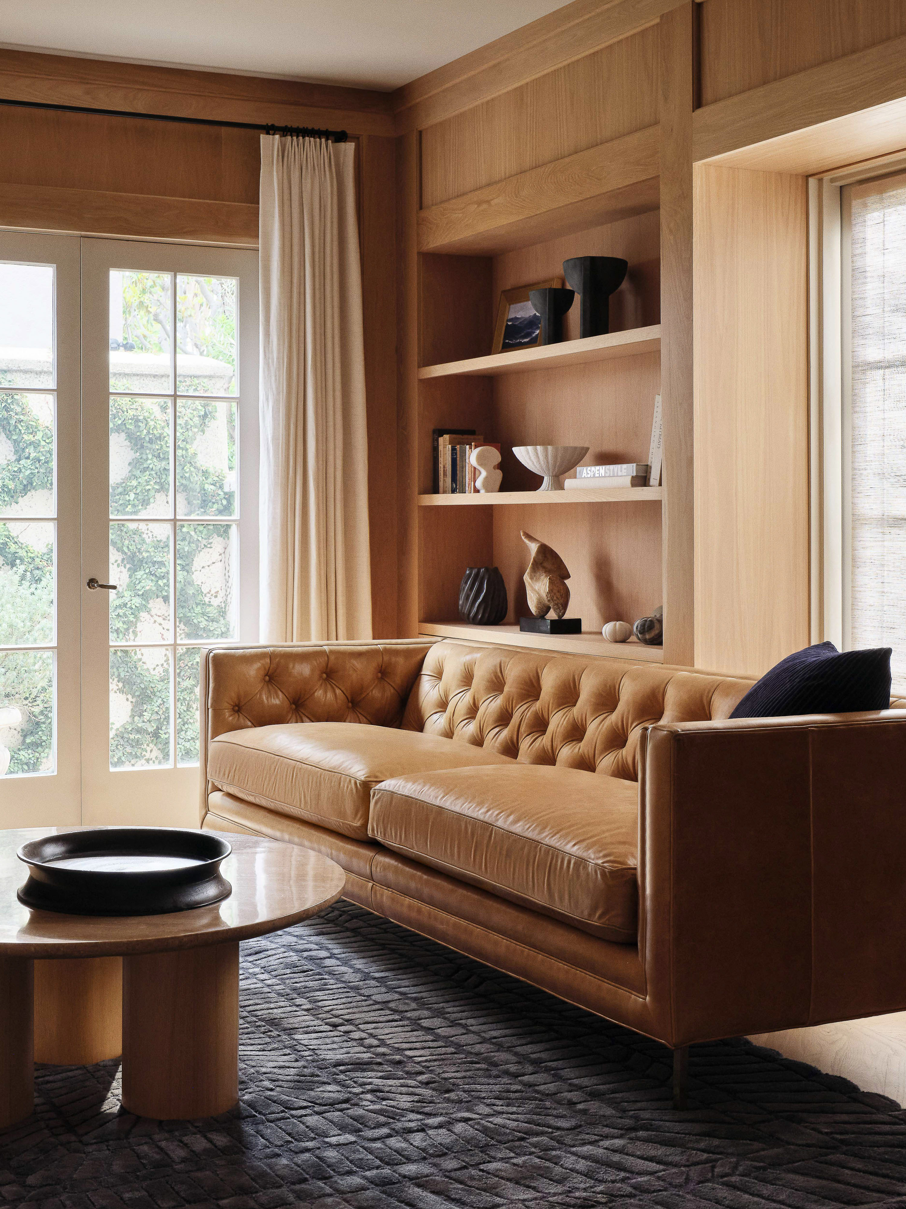 Living room interior featuring a tan leather sofa, round coffee table, patterned rug, and wooden shelving with decorative objects.