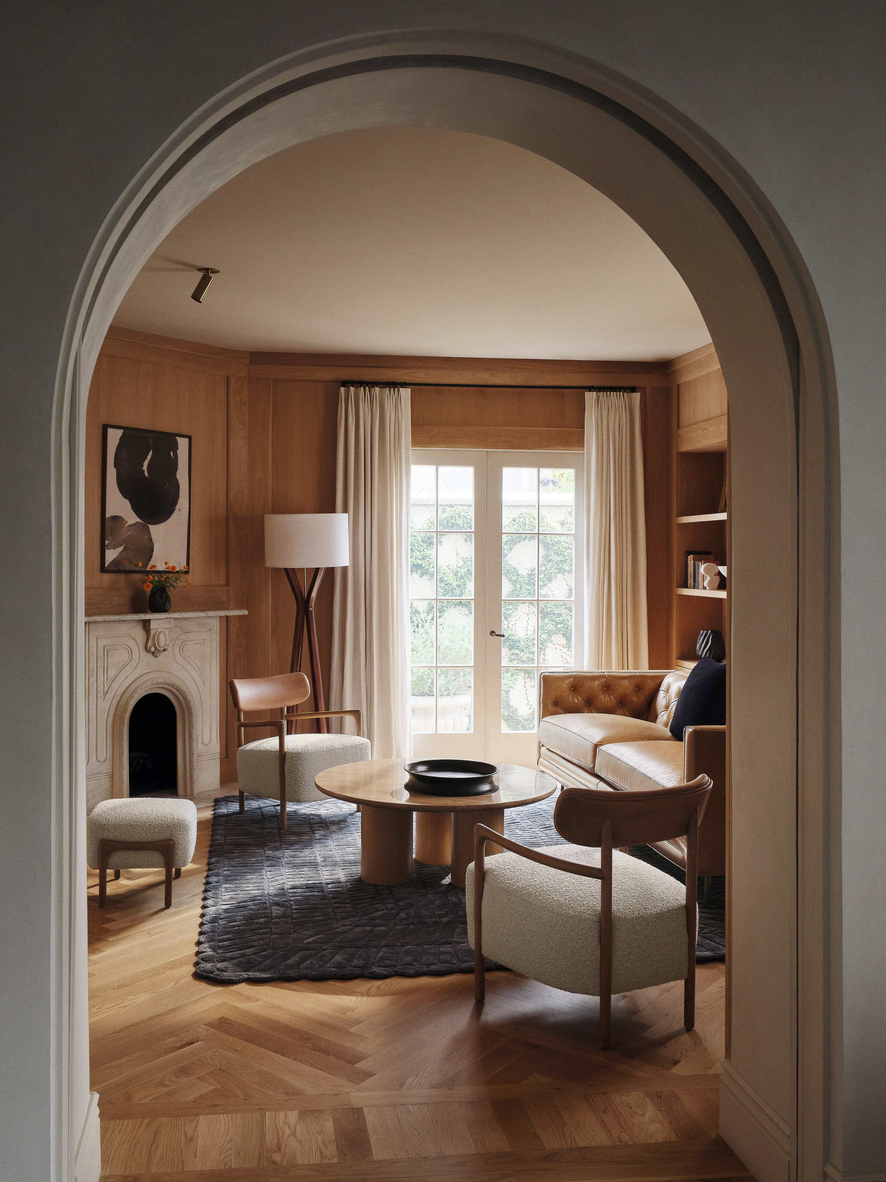 Interior view of a living room with two armchairs, a round coffee table, a sofa, and a fireplace, seen through an archway entrance.
