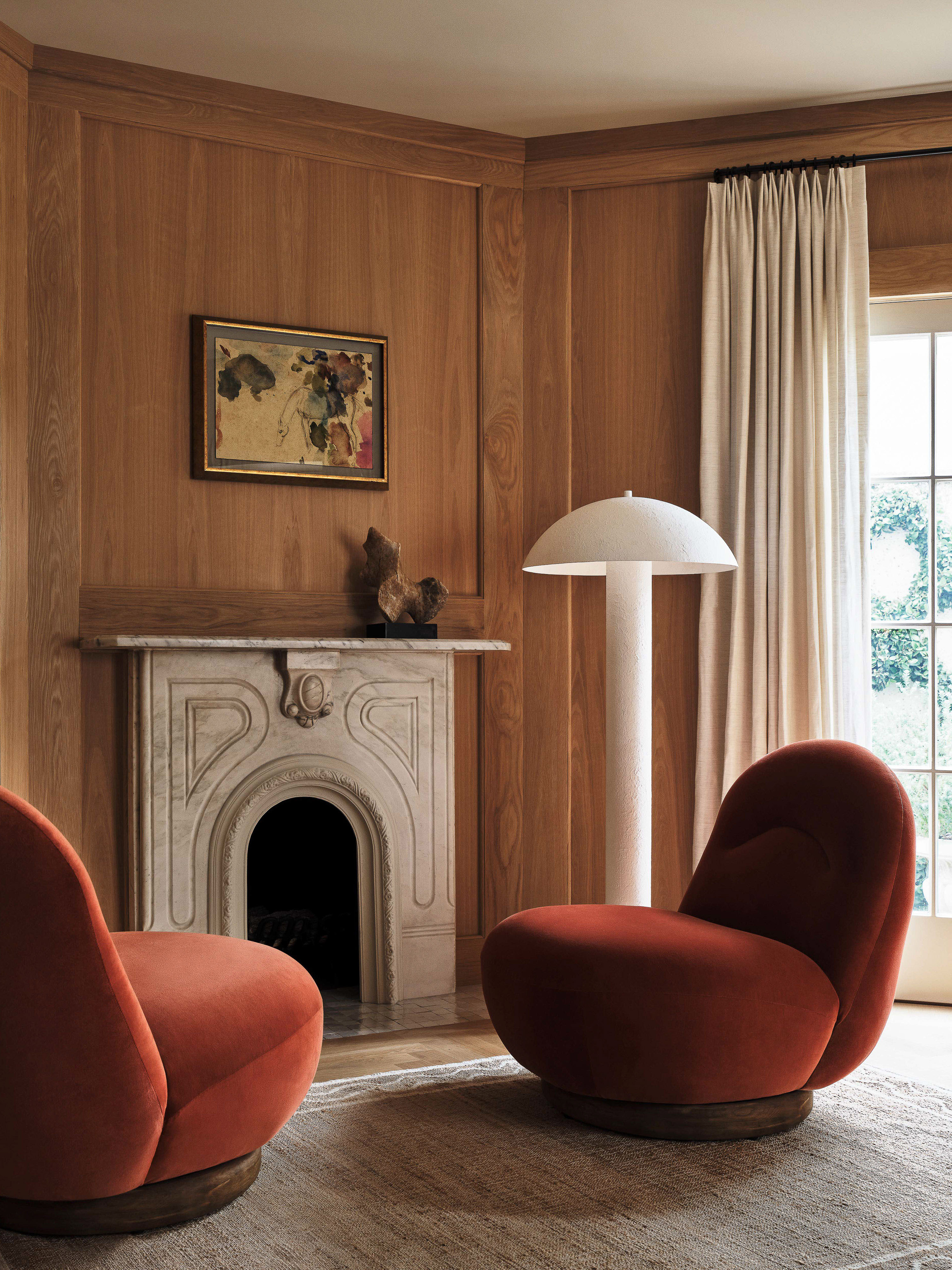 Interior view with two red chairs facing fireplace, stone mantel, wall art, and mushroom lamp in wood-paneled living room.